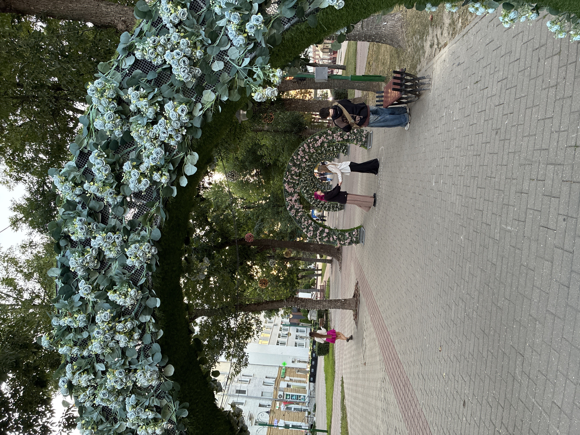 The image depicts an outdoor scene in a city park in Mahilyow, Belarus. A large, decorative archway adorned with artificial greenery and blue flowers frames the foreground. In the middle ground, two individuals are standing close together, possibly interacting or posing for a photo, with one wearing a pink hijab and the other a white top and dark skirt. To the right, another person is standing, looking towards the other two. A young woman with long, dark hair and a pink skirt is walking away from the camera on a brick-paved pathway. In the background, there are trees, buildings, and streetlights, suggesting an urban environment. The overall impression is of a pleasant, possibly festive, outdoor gathering.