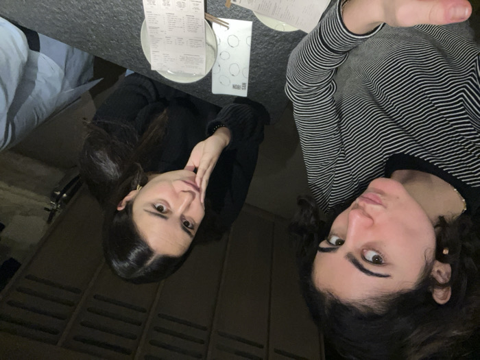 Two women pose for a selfie at a restaurant table. One woman, on the left, wears a black sweater, her face tilted with her hand near her chin. The second woman, on the right, wears a striped shirt. A menu, a plate, and utensils are on the table. A blue object partially visible in the upper left corner. The angle is slightly tilted.