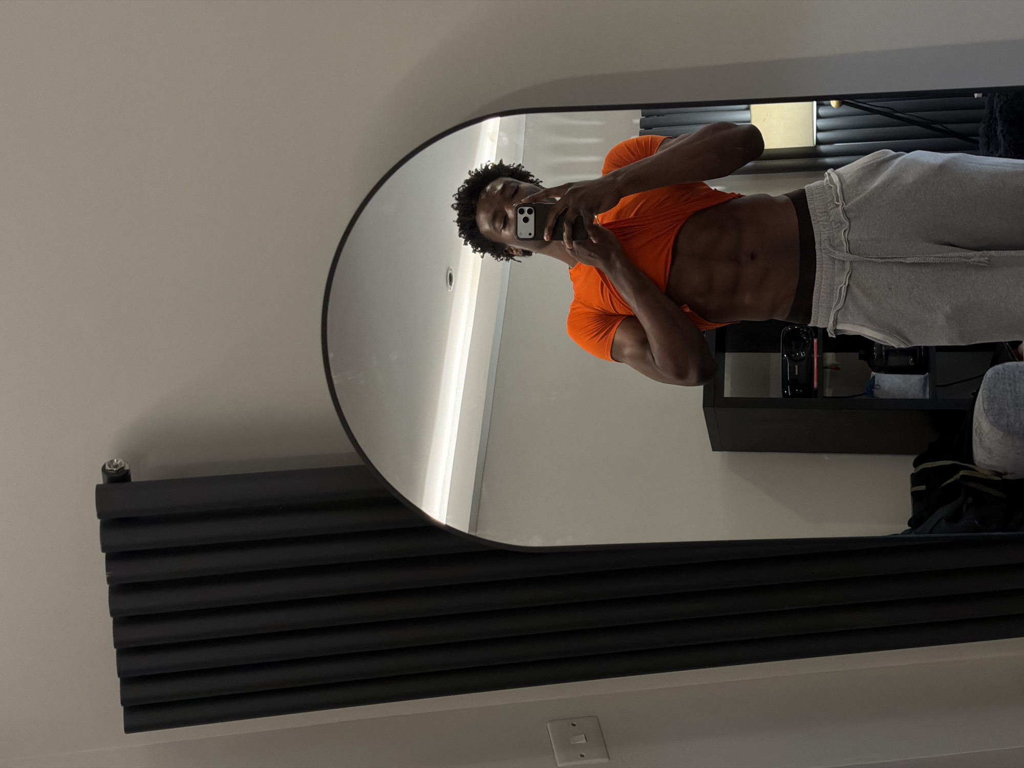 A well-lit interior scene depicts a man taking a mirror selfie. The man is shirtless, wearing an orange top and grey sweatpants. He has dark hair and holds a smartphone in front of his face, obscuring it partially. His toned abdomen is visible.
The selfie is taken in a large, rounded-edge mirror hanging on a light beige wall. To the left of the mirror, a modern black radiator is mounted on the wall. Reflected in the mirror is a shelf unit with various items, including a black device, books, and a partially visible lamp with a white shade. A dark object resembling luggage or a bag is visible near the base of the shelf unit. A light switch is situated beneath the radiator on the wall.
The environment appears clean and modern, suggesting a domestic setting. While it's impossible to confirm the specific location within London from this image alone, the interior design style and light colors are characteristic of contemporary London apartments.