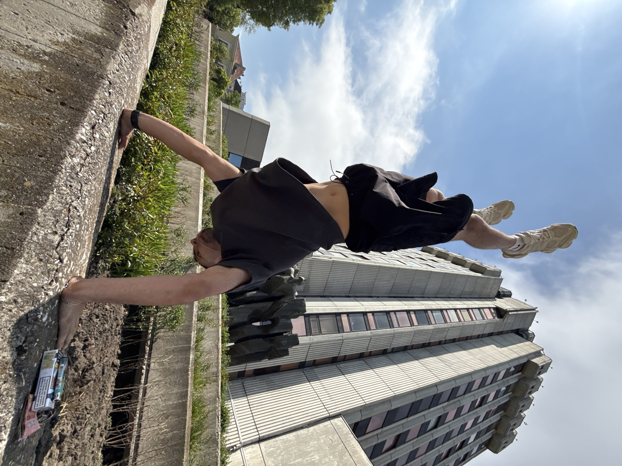 A man is performing a handstand on a low concrete wall in Varna, Bulgaria. The building in the background is the Interhotel Cherno More. The man is wearing dark shorts, a dark t-shirt, and light-colored sneakers. He is holding onto the wall with both hands, and his body is extended horizontally. There are cigarettes and cash in the space below his hands. The sky is blue with scattered clouds. Vegetation grows along the wall.