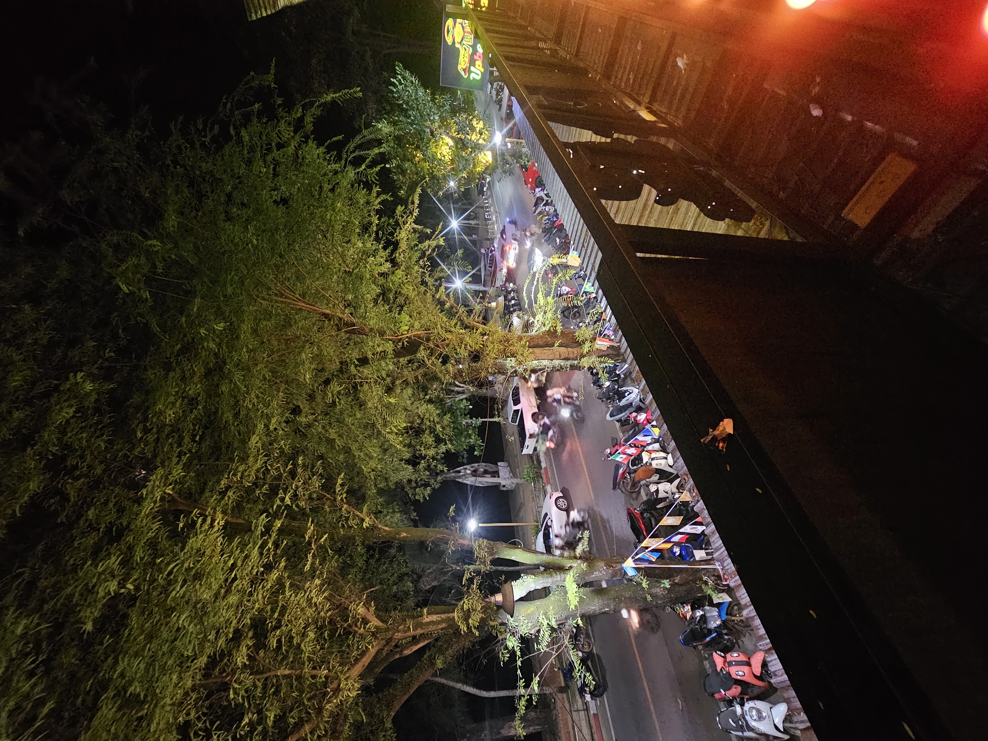 This image captures a nighttime scene in Ban Klong Haeng, Thailand. The perspective is from a slightly elevated vantage point, looking down at a street lined with trees and parked motorcycles. A building with a corrugated metal roof is visible on the right, with warm, orange light emanating from it.
The street is illuminated by streetlights and the lights from the building, casting a yellow and orange glow on the surroundings. Several trees with dense green foliage are prominent in the foreground and along the street, their leaves catching the light. A variety of motorcycles are parked along the side of the road and on a sidewalk, some with colorful decorative flags. A few cars are also present on the street.
People can be seen on the sidewalk and near the building, though their features are not clearly discernible due to the nighttime lighting and distance. The activity appears to be people gathering or passing by, likely at a restaurant or other establishment. The overall atmosphere suggests a lively, yet relaxed, evening setting.
There is a neon sign visible above the building, with text that is partially legible. The sign appears to be green and yellow, with stylized lettering and graphics, possibly indicating a food establishment. The time of day is clearly night. The weather appears clear, as evidenced by the absence of rain or fog. The presence of numerous motorcycles and the type of buildings suggest an urban or semi-urban environment in Southeast Asia.