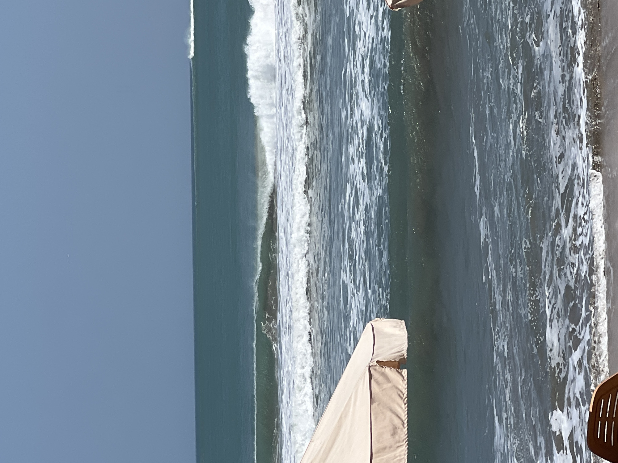 The image depicts a serene beach scene in Kuta, Indonesia, with a clear blue sky overhead transitioning to a turquoise ocean. Gentle waves roll towards the shore, creating a white foamy surf. In the foreground, a portion of a beige beach umbrella and a brown plastic chair are visible. The composition suggests a tranquil day at the beach, with the vast ocean as the dominant element.