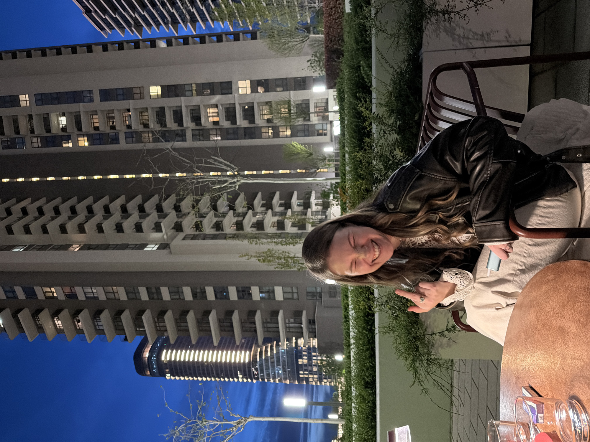 A horizontally-oriented, medium shot captures a smiling Caucasian woman seated at a table on an outdoor patio, with buildings and a darkening sky visible in the background. The woman, turned slightly to the left, is wearing a black leather jacket over a light-colored top with lace detailing. She is making a hand gesture with her left hand, displaying a "peace" or "victory" sign.
She sits on a chair with a metal frame and horizontal slats. A round, wooden-like table is to her right, with glassware visible near the bottom edge. The patio is lined with green bushes and vegetation.
Behind the patio, several modern, high-rise buildings dominate the skyline. The buildings are characterized by multiple windows and layered architectural elements. The sky transitions from a deep blue at the top to a lighter shade near the horizon.
Given the architecture, the environment, and visual cues, the location is identified as Belgrade, Serbia. The presence of modern high-rise buildings suggests a newer or redeveloped area of the city.