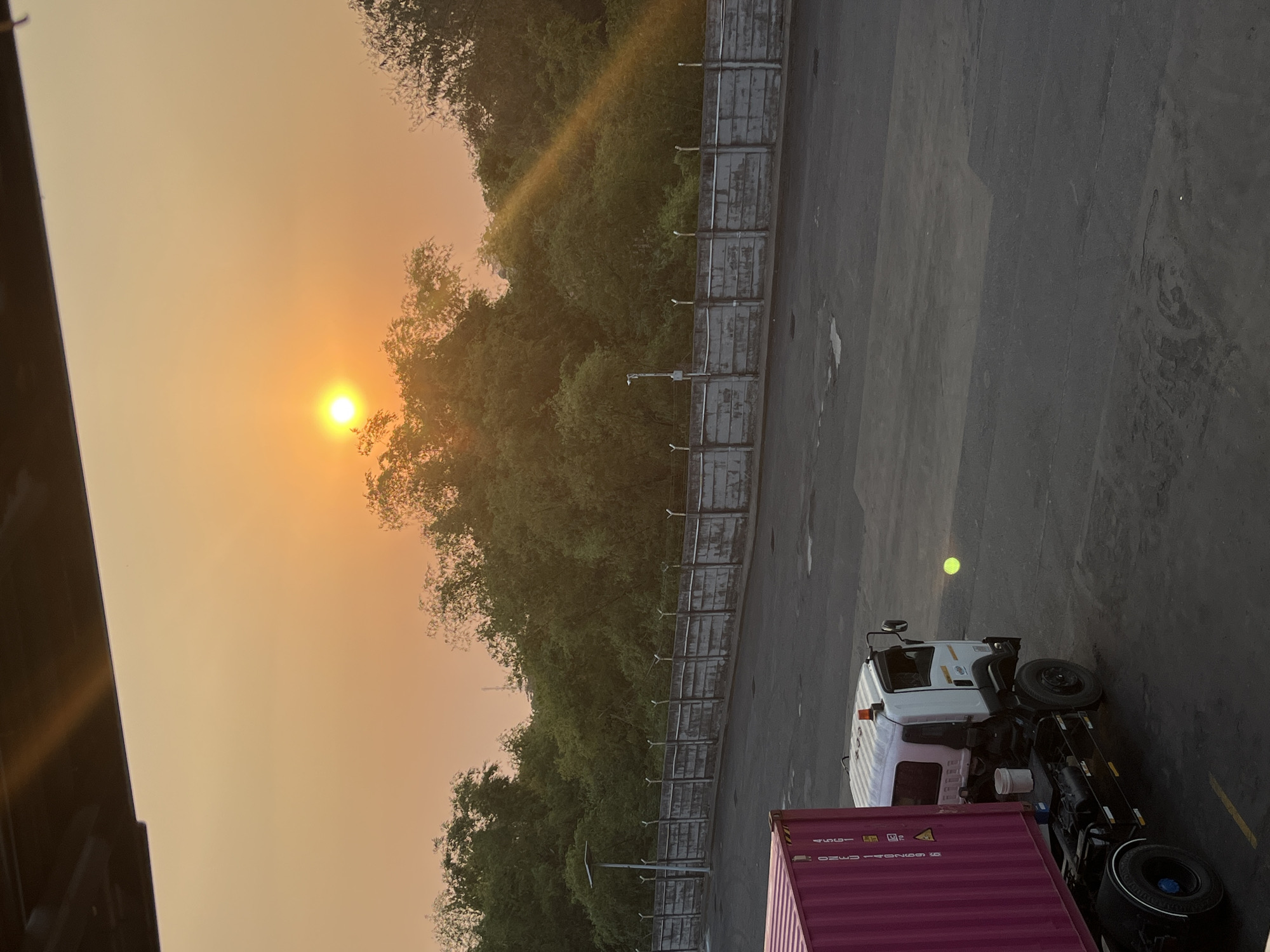 The image shows a sunset at a port in Pelang, Indonesia. The sun is setting in the distance, casting a warm orange glow over the sky and trees. In the foreground, a truck with a pink shipping container is parked on an asphalt surface. A concrete wall with a barbed wire fence runs along the middle ground, separating the port area from the trees behind it. The scene is quiet and peaceful, with no people visible.