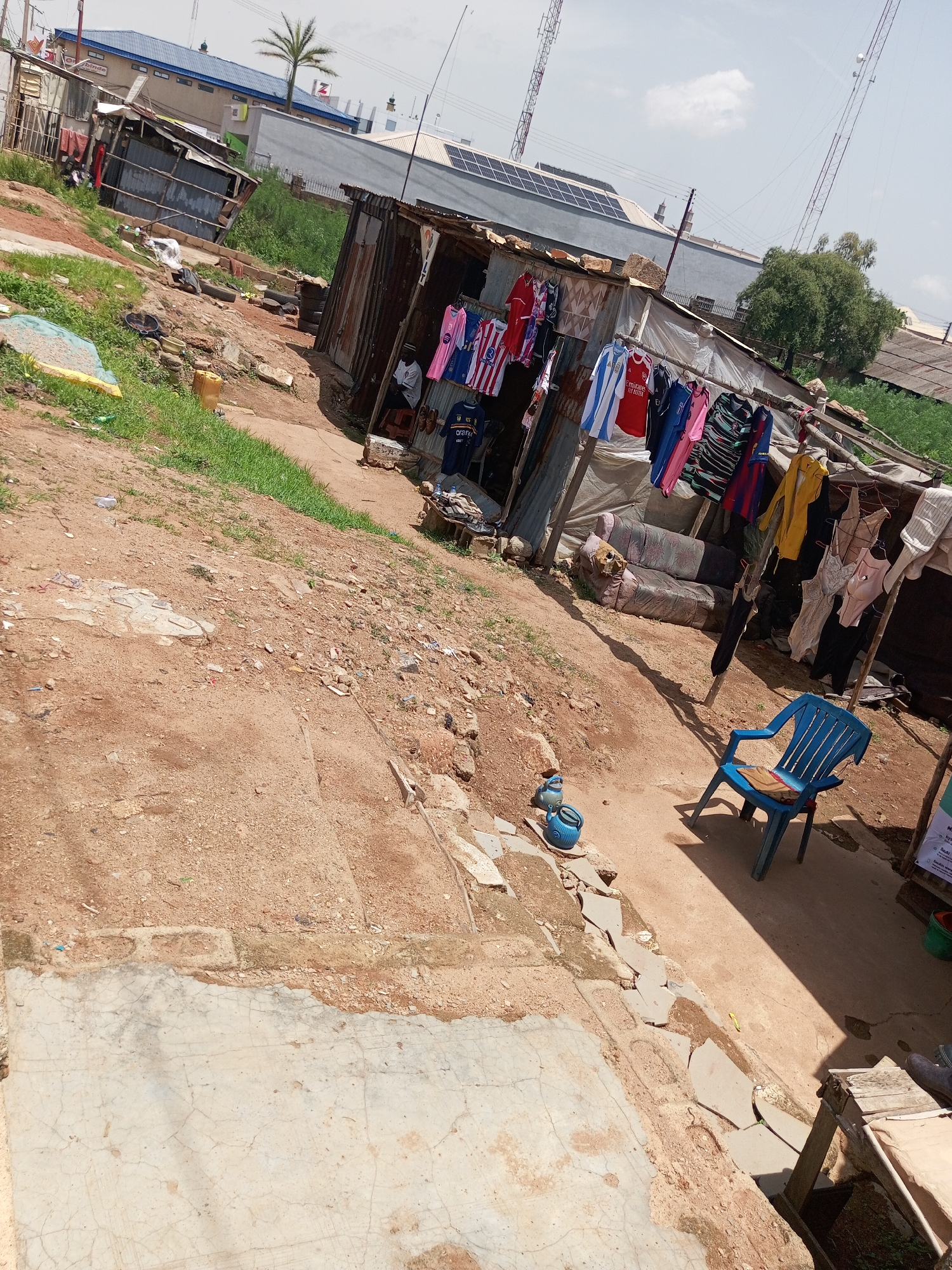 This is an outdoor, eye-level medium shot of a street market scene in Bukuru, Nigeria. The setting is rustic and appears to be a developing area, with a mix of corrugated metal structures and some more modern buildings in the background. The ground is a mix of dirt and cracked concrete.
In the foreground and midground, the scene is dominated by stalls selling clothing, primarily what appear to be football jerseys. Several jerseys are hanging on a clothesline, displaying various colors and team logos. Some are neatly folded on display. A young person, likely a vendor, is seated in one of the stalls, observing the surroundings. Nearby, a bright blue plastic chair sits empty, with a brown object, possibly a book or notebook, resting on its seat. Two blue ceramic pots are placed on the ground next to the chair.
In the background, a multi-story building with a blue roof is visible, featuring solar panels on its upper level. Behind this, taller structures resembling communication towers rise into the sky. Lush green vegetation can be seen in the distance, suggesting a warm climate. The sky is bright and appears to be mostly clear, indicating a daytime setting, likely late morning or early afternoon, with the sun casting distinct shadows.
A sign with text is partially visible on the right side of the image, but the text is too blurry to read. The overall impression is one of a vibrant but simple marketplace, possibly experiencing a period of development given the contrast between the older structures and the newer buildings in the background. The scene conveys a sense of everyday life in this Nigerian location.