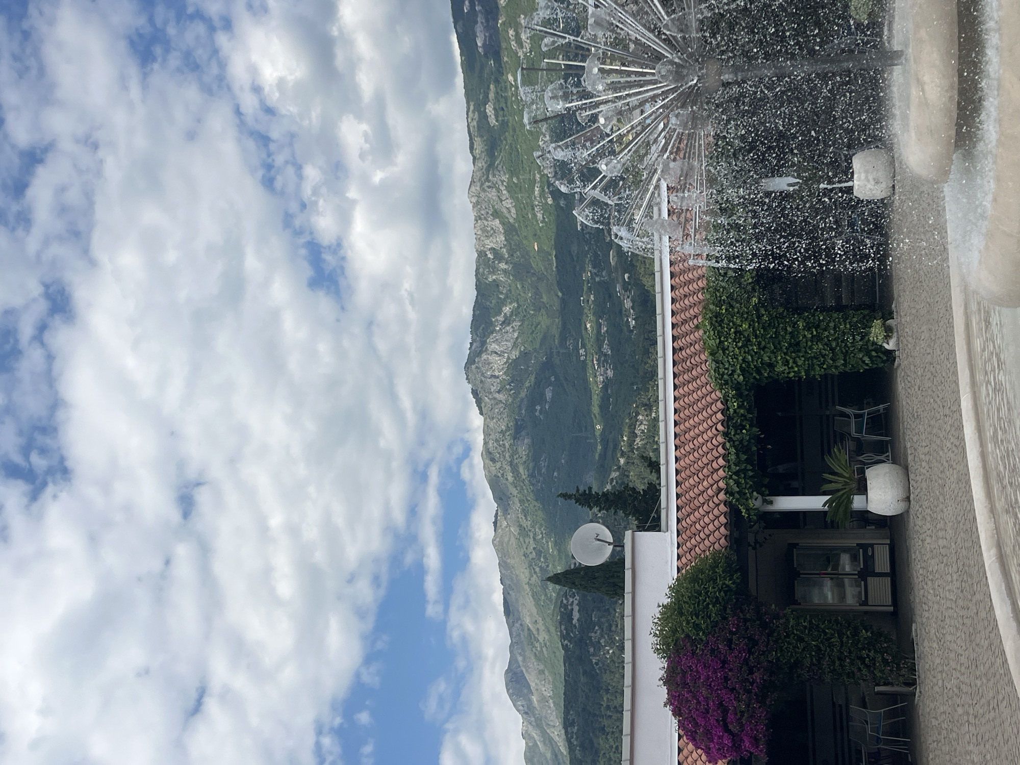 The image presents an outdoor scene in Budva, Montenegro. The view is partially obscured by a fountain with a spherical spray structure in the foreground, casting water droplets across the scene.
Behind the fountain, a building with a red-tiled roof and a light-colored exterior is visible. Green vegetation and purple flowers adorn parts of the building. Through an open window, there's a view of a terrace with chairs and potted plants. A satellite dish is mounted on the side of the building.
In the background, a mountain range dominates the landscape, partially obscured by clouds. The sky features a mix of blue patches and white clouds.