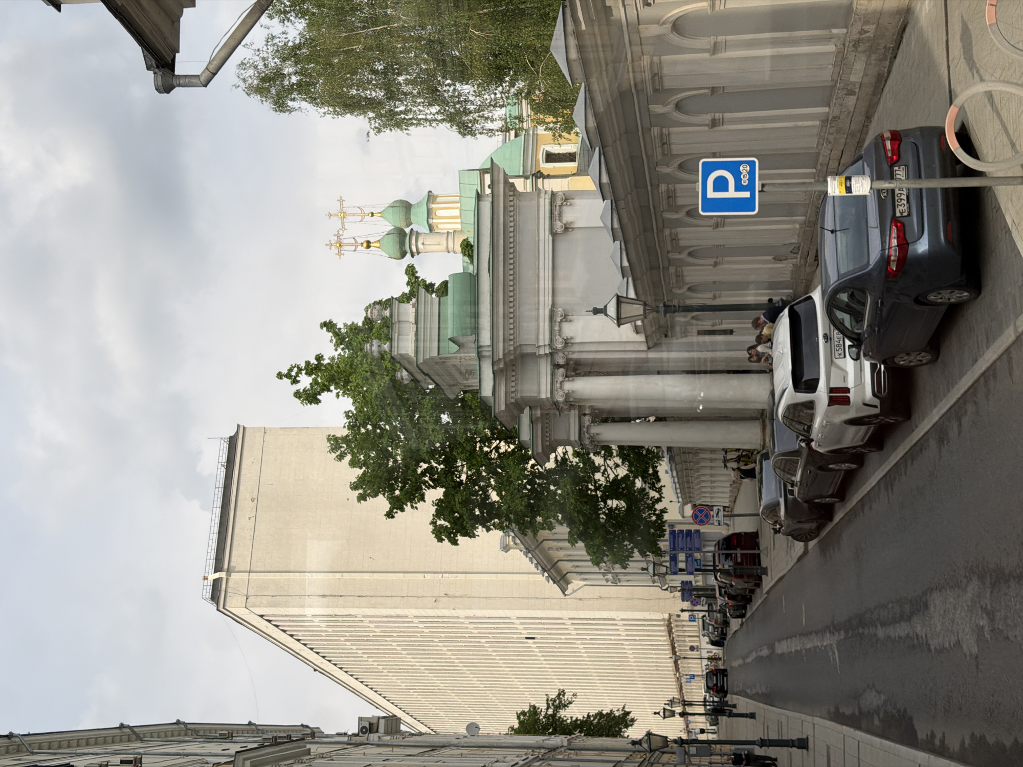 The image depicts a street scene in an urban area. A tall, off-white building with a grid-like facade is on the left side of the frame. To its right, a smaller building with classical architectural details and green domes is visible. A line of parked cars is on the right side of the street, with a blue parking sign near the closest car. A wet road runs between the buildings and the cars. Street lamps line the street. People are gathered near the cars. The sky is overcast. The location is Unknown, Unknown.