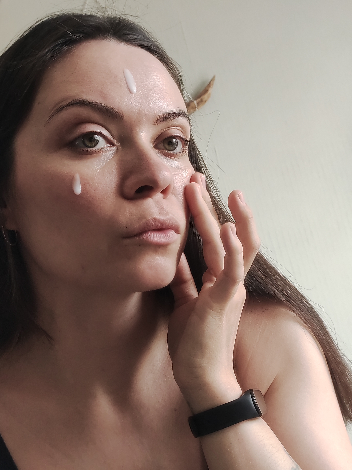 A woman with light skin and long, dark brown hair is applying cream to her face. There is a drop of cream on her cheek and a dollop on her forehead. She is looking at the camera while touching her right cheek with her right hand. A black fitness tracker is on her wrist. The background is a light beige wall. The exact location in the city of Chaikovsky, Russia, cannot be determined from the image.