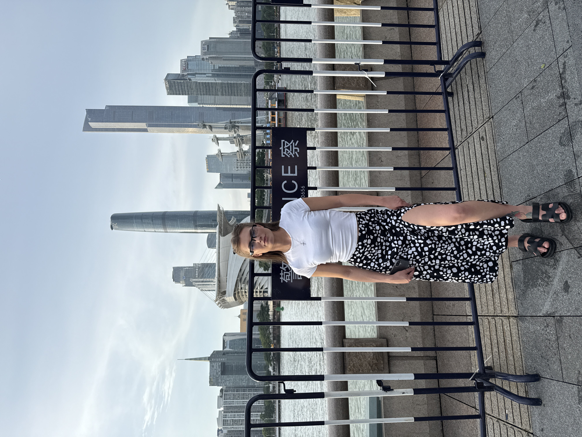 A medium shot captures a woman with blonde hair wearing glasses, a white t-shirt, and a black and white spotted skirt with a high slit, standing on a paved walkway in front of a body of water. She is looking at the camera, holding a dark-colored smartphone in her left hand. Behind her, a black and white striped metal barricade separates the walkway from the water. In the background, a cityscape with numerous tall buildings, including the distinctive IFC tower, is visible against a pale sky. The scene appears to be a waterfront promenade in the Haizhu District of Guangzhou, China, suggested by the architecture and signage which appears to include Chinese characters.