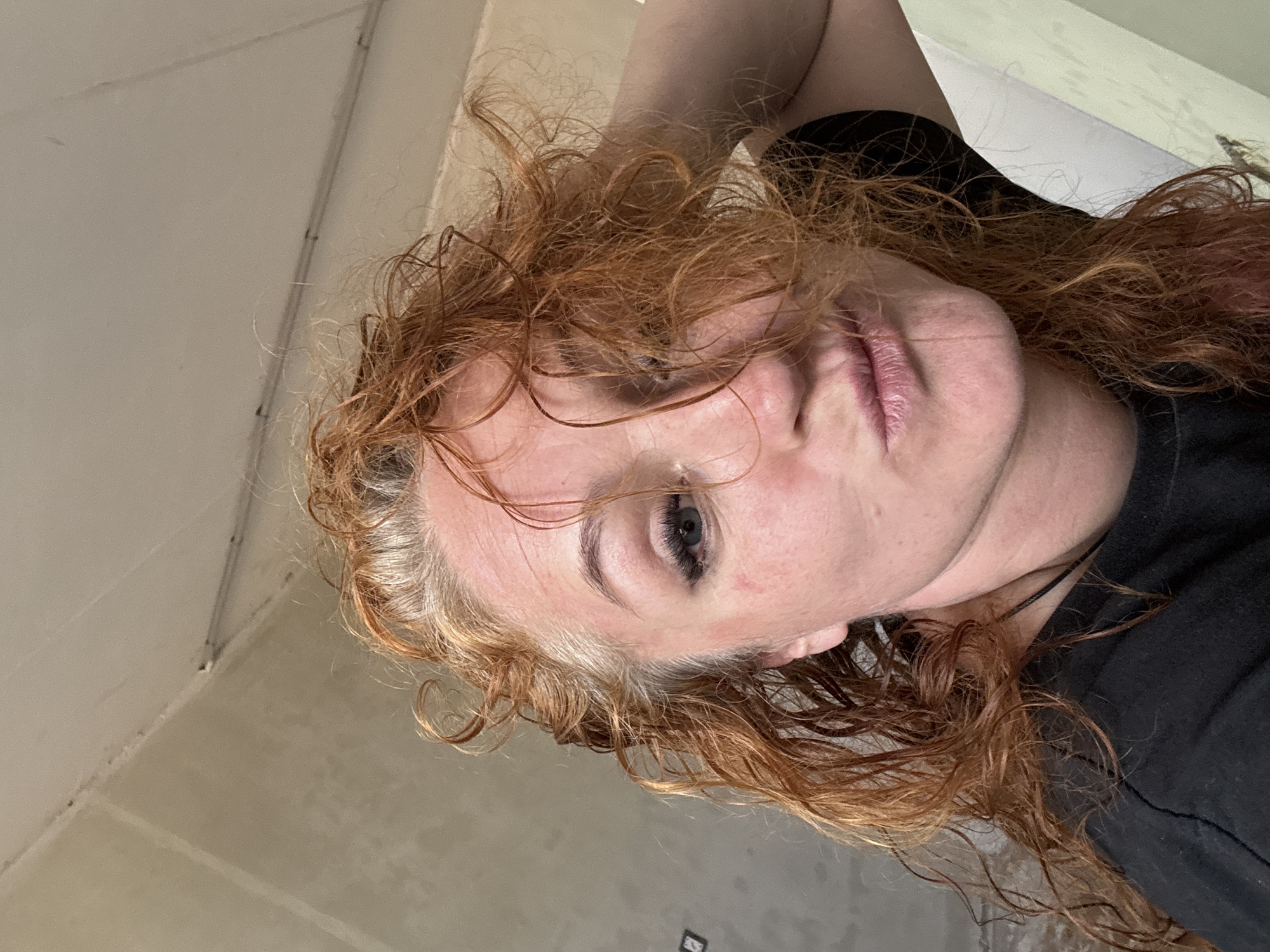 A selfie image captures a person with auburn, wavy hair, likely indoors against a pale beige wall. The subject has fair skin, one visible blue eye, and is wearing a black top. Details of the environment are minimal, showing just a glimpse of the interior wall. The image is angled, so the ceiling is visible in the upper left. There are no overt indications this photo was taken in Chaikovsky, Russia.