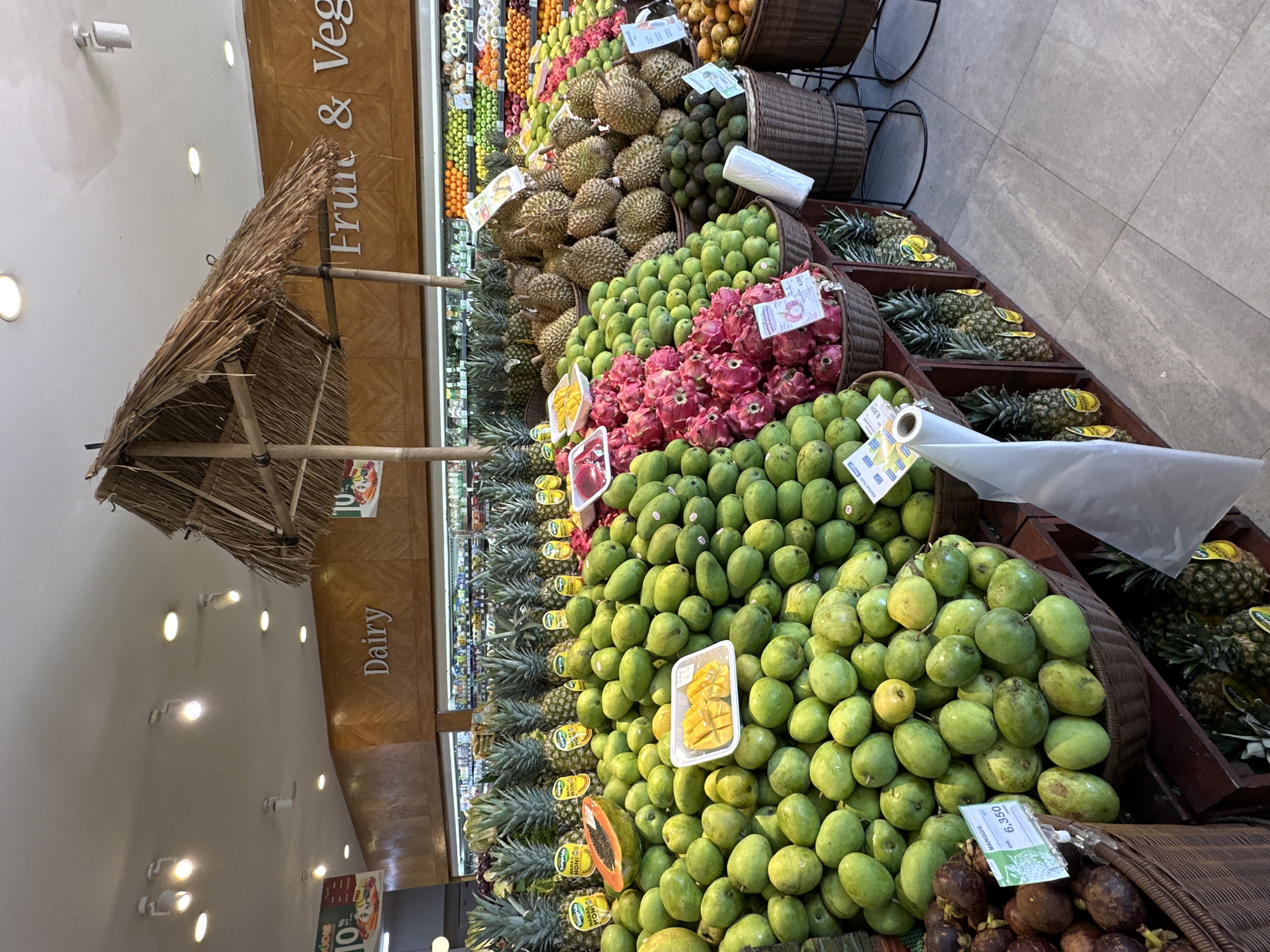 The image shows a colorful display of various fruits in a market or grocery store in North Kuta, Indonesia. The foreground features arrangements of green mangos, pineapples, dragon fruits, durians, and other tropical fruits in baskets and tiered displays. There is a thatched hut-like structure above the fruit display, potentially part of the store's decor. Store signage for "Fruit & Veg" and "Dairy" can be seen in the background. The lighting suggests an indoor environment.