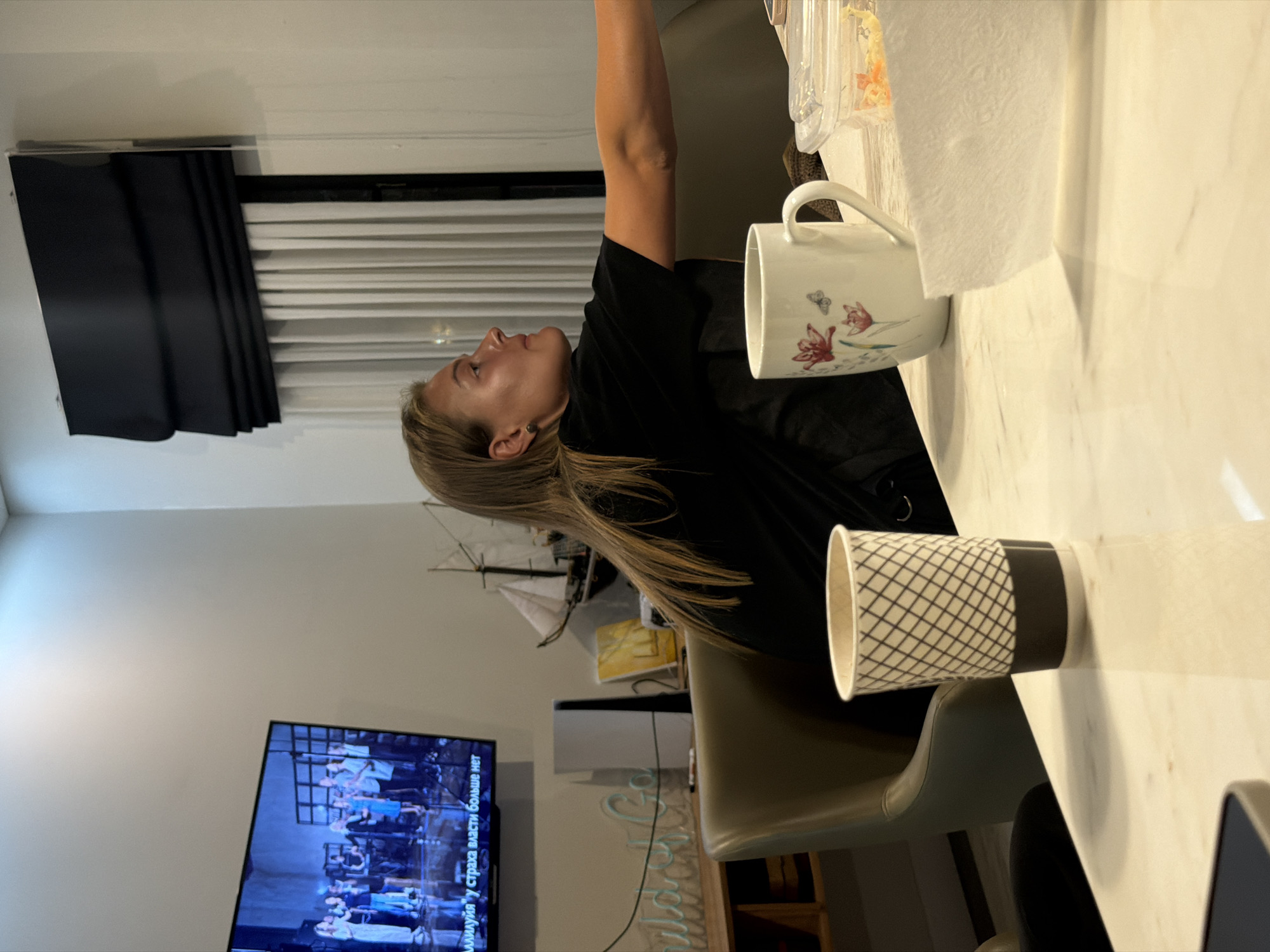 The image shows the interior of a room, potentially in a residence. A woman with blonde hair is seated at a table. She is wearing a black shirt. On the table are two mugs, one white with floral designs and one with a black and white pattern, along with some containers and paper towels. In the background, there's a television displaying an image. A model ship is visible behind the woman. The setting could be Kerobokan, Indonesia, but this cannot be confirmed from the image alone.