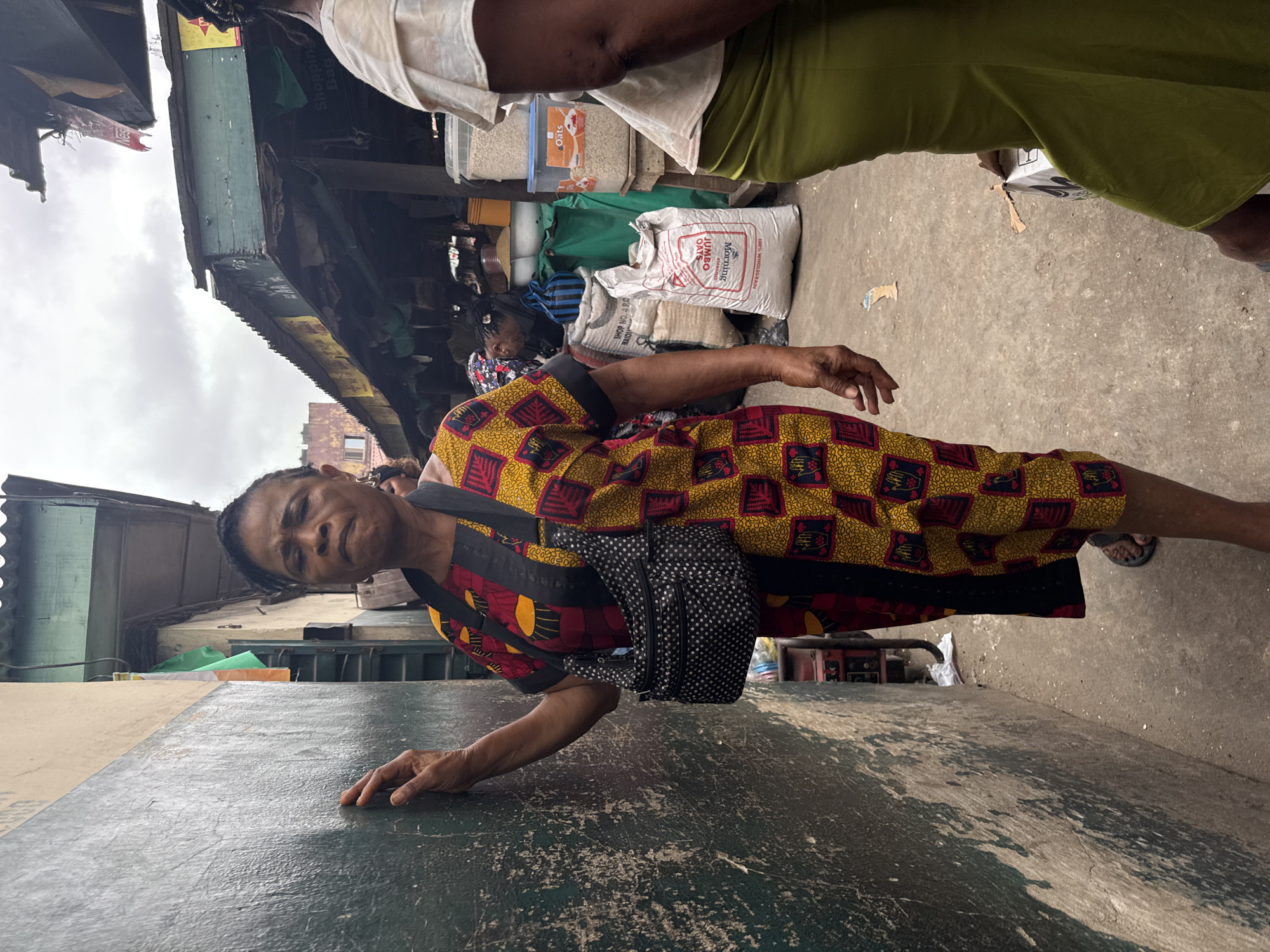 A medium shot captures an African woman in Lagos, Nigeria, standing in a marketplace. The woman is positioned in the lower center of the frame, leaning against a textured, dark green wall on the left. She is wearing a vibrant yellow and red patterned dress and a black and white polka dot crossbody bag. Her left hand rests on the wall, and her right arm is bent at the elbow, with her hand near her chest. She looks directly at the camera with a neutral expression.
Behind her, the marketplace unfolds. Stalls with corrugated metal roofs are visible, adorned with various goods and advertisements. Several large sacks of grain or produce are stacked on the ground, some with visible labels. A white bag with red lettering reads "JUMBO Oats." To the right, the back of another woman wearing a light-colored top and a lime green skirt is partially visible. The ground is a concrete surface, showing some debris and unevenness. The sky above is overcast. The overall impression is one of a busy, everyday scene in a Nigerian marketplace.