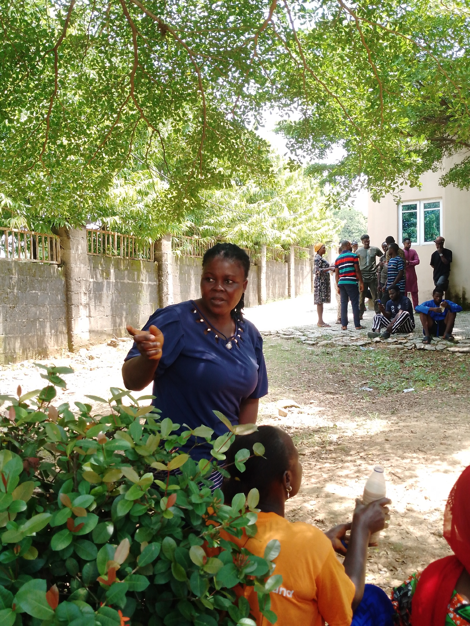 In Akwanga, Nigeria, under the dappled shade of a tree, a group of people are gathered outdoors. In the foreground, a woman wearing a navy blue top is gesturing with her right hand as she speaks. Partially visible in the lower left is a dense green bush. Behind her, a group of individuals are standing and sitting, some conversing. A building with a window is visible in the background, with a fence running along the side. The ground is covered with a mix of dirt, stones, and patches of grass.