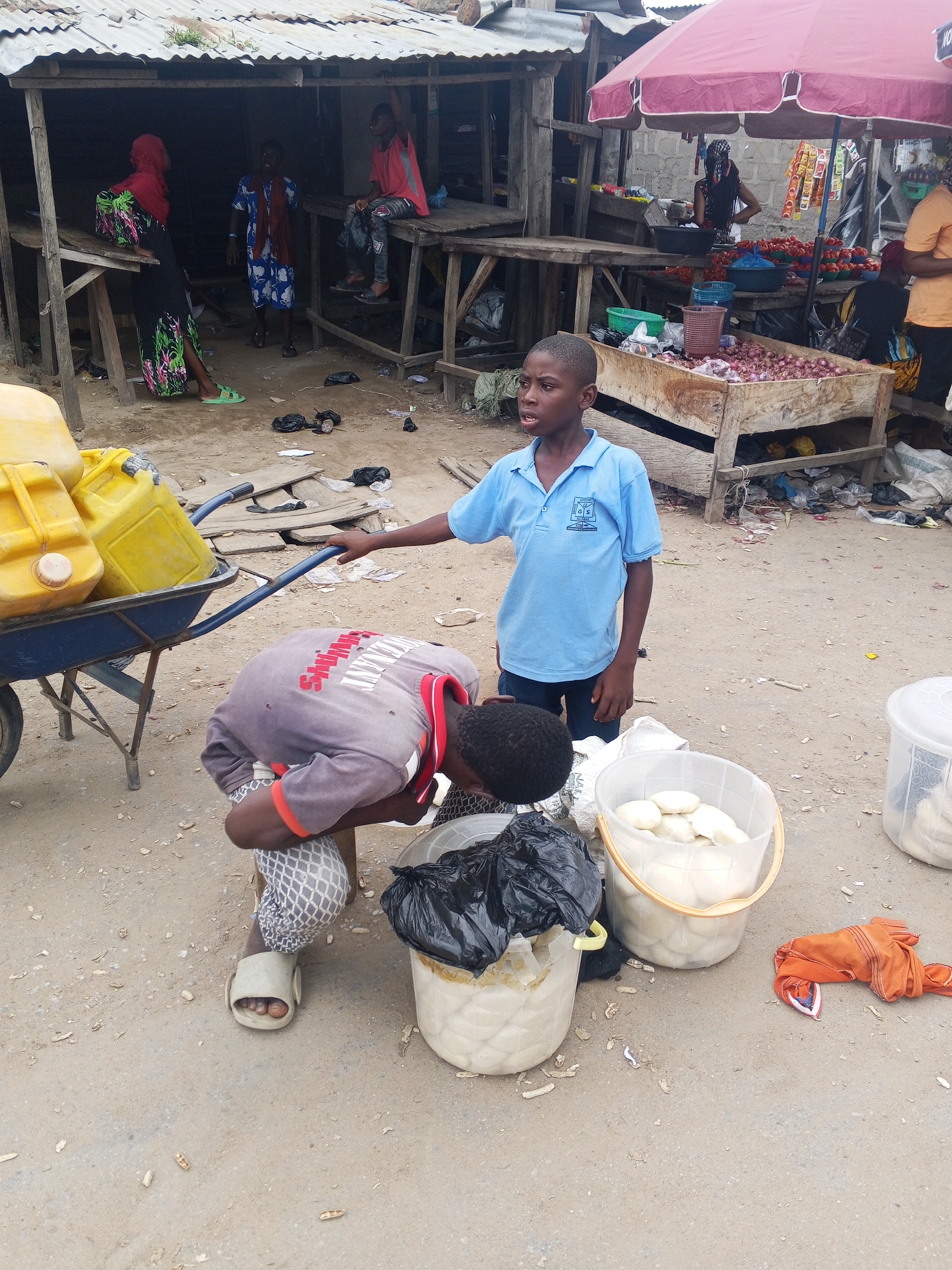The image depicts a scene at an outdoor market, possibly in Usunwe, Nigeria. In the foreground, two young boys are engaged in activity around a blue wheelbarrow loaded with yellow jerrycans. One boy, wearing a blue polo shirt with a school crest, stands looking towards the right. The other boy is bent over, seemingly examining or interacting with items near the wheelbarrow. Beside them, several large white buckets are present; one contains white, round food items, another is filled with a similar food substance and partially covered with a black plastic bag, and a third white bucket is visible to the right. An orange garment lies on the ground to the right of the buckets. In the background, under a corrugated metal roof structure, several people are visible, including individuals wearing a red hijab and a blue patterned dress. The market stalls appear rustic, with wooden structures and an array of goods, including red tomatoes and onions in a wooden crate under a pink umbrella. The ground is dusty earth, with scattered debris and what appear to be peanut shells. The overall environment suggests a busy, informal marketplace.