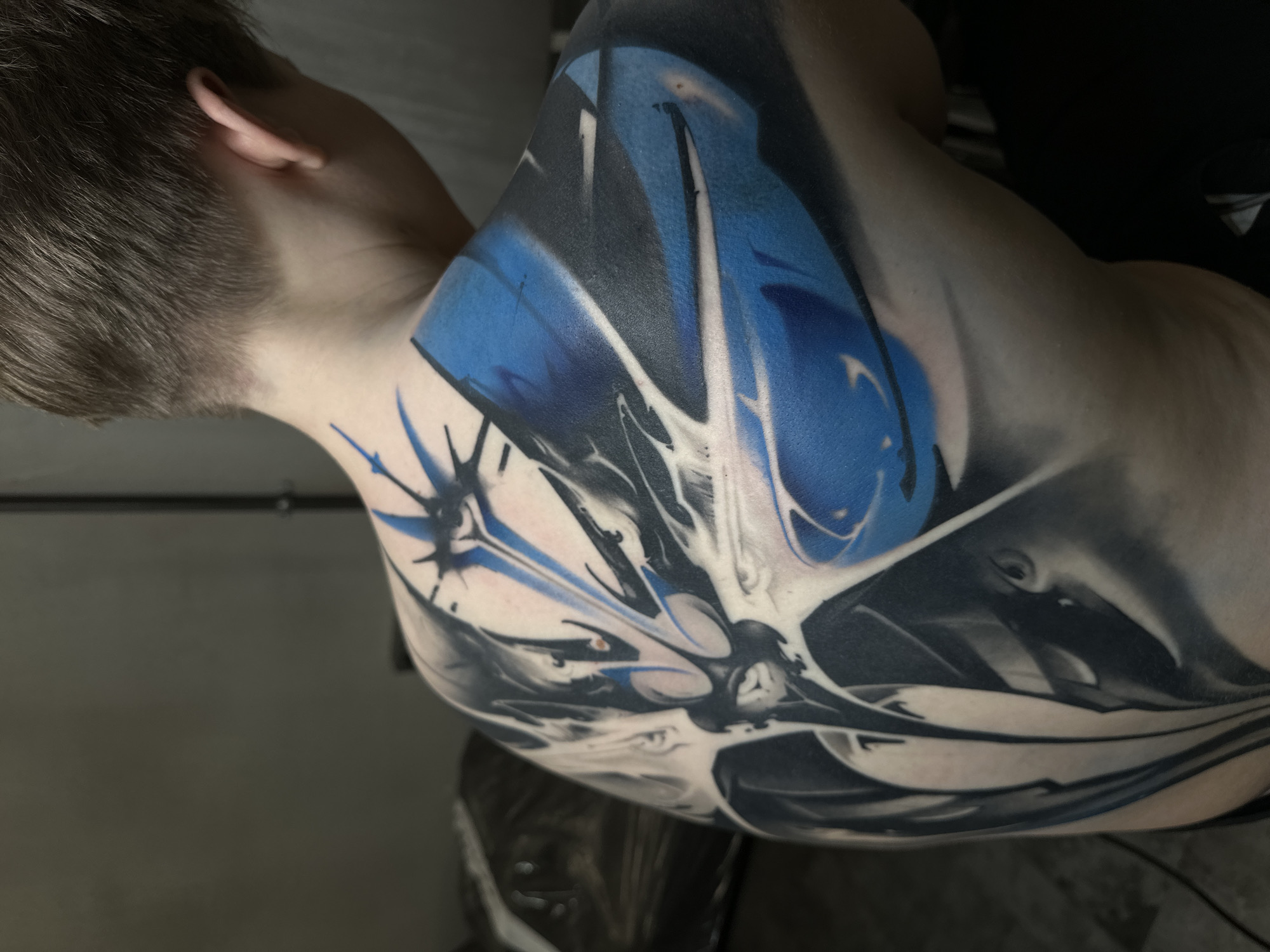 A close-up shot focuses on the back and neck of a person, likely a young adult male, featuring a large, abstract tattoo. The tattoo dominates the view, covering much of the shoulder and upper back. The tattoo incorporates a palette of blue, black, and white, with a design that appears dynamic and possibly futuristic or biomechanical. The person's hair is short and neatly cut. The background is nondescript, consisting of a dark wall and possibly part of a chair or object in a muted tone. There are no clear indications of the location being Moscow or Russia in the image.