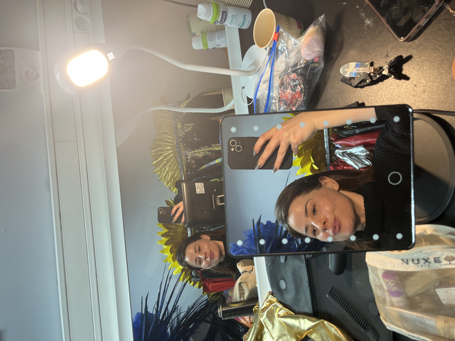 The image showcases a cluttered dressing table. The main subject is a woman taking a selfie in a light-up mirror. Various cosmetic items, including containers labeled "Sanex Zero," sit near the mirror. A desk lamp illuminates the scene. The reflection in the mirror reveals a wall with a flamboyant costume or artwork featuring feathers and metallic accents. A plastic bag containing makeup sponges, hair clips, and a Nuxe bag are also visible.