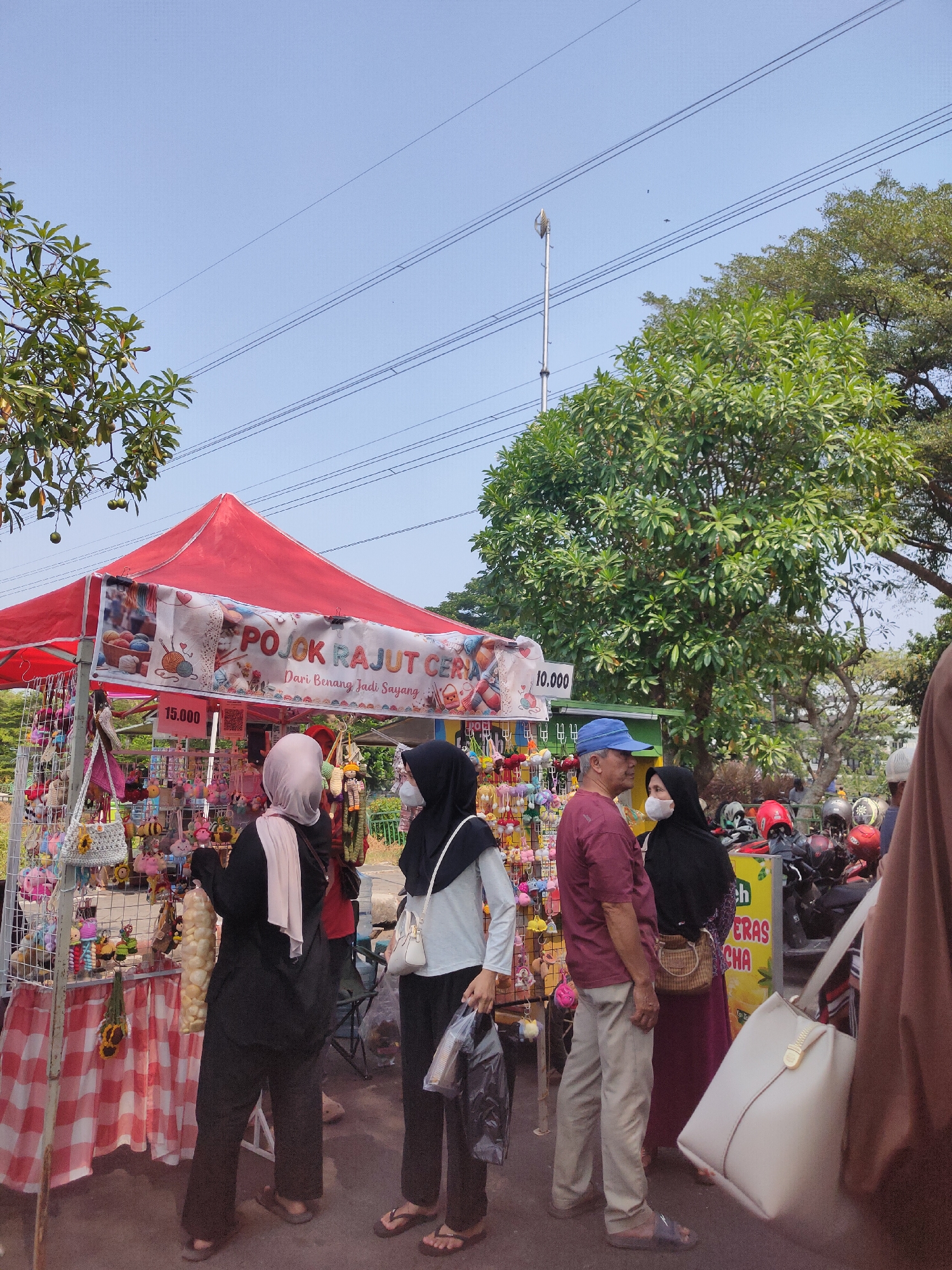 The image shows an outdoor market stall in Ngasem, Indonesia, on a sunny day. The stall is decorated with a red canopy and a banner that reads "POJOK RAJUT CERIA" and "Dari Benang Jadi Sayang," indicating it sells knitted items. Various small knitted toys and keychains are displayed on racks.
Several people are present at the stall. Two women wearing hijabs are browsing the merchandise. One woman in a black hijab and light blue top is holding a plastic bag. Another woman in a black hijab and a black outer garment is looking at the items. A man in a maroon t-shirt and blue cap is standing near them, facing away from the camera. Behind the stall, there are motorcycles parked and more people in the background. The ground appears to be asphalt. Trees and power lines are visible in the background, suggesting an urban or suburban setting. The overall atmosphere seems to be that of a local market or fair.