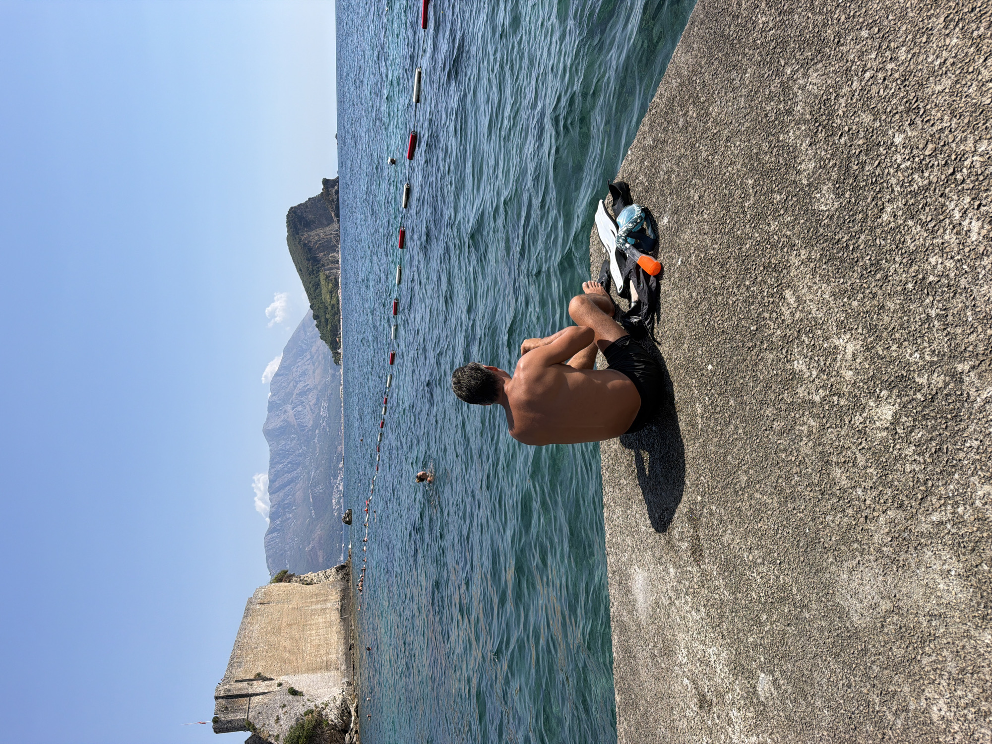 A man sits on a concrete pier overlooking the Adriatic Sea in Budva, Montenegro. He is facing away from the camera, with his back to the viewer. The man is shirtless, wearing black swim trunks, and has short brown hair. Next to him on the pier are a black bag, fins, and snorkeling gear, including a mask and an orange snorkel. The sea is a vibrant turquoise color, with visible waves. In the background, to the left, is the historic Old Town of Budva, with its stone fortifications perched on a rocky outcrop. Further in the distance, a green, mountainous island rises from the sea, and more mountains are visible on the mainland. A line of buoys demarcates a swimming area in the water. Several people are swimming in the sea, and a few are visible on the beach in the distance. The sky is clear and blue, indicating a sunny day.
