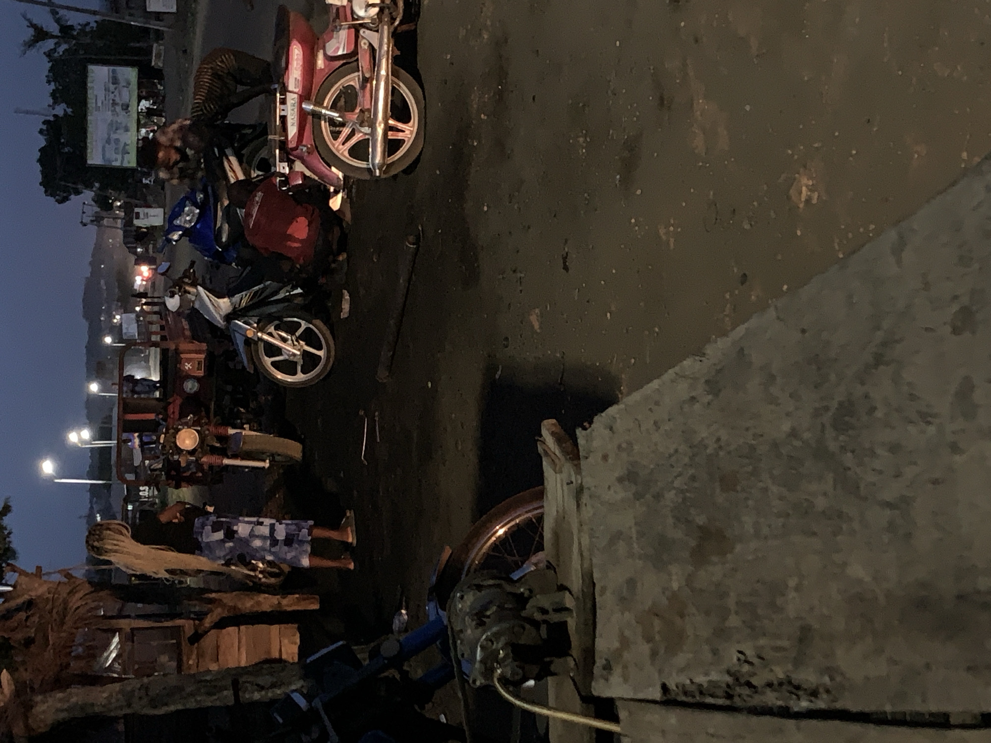 The image shows a street scene in Gbeleko, Nigeria at dusk. There are several motorcycles parked on the street. People are visible, some appearing to be working on the motorcycles. Streetlights illuminate the scene. A building structure is visible in the foreground.