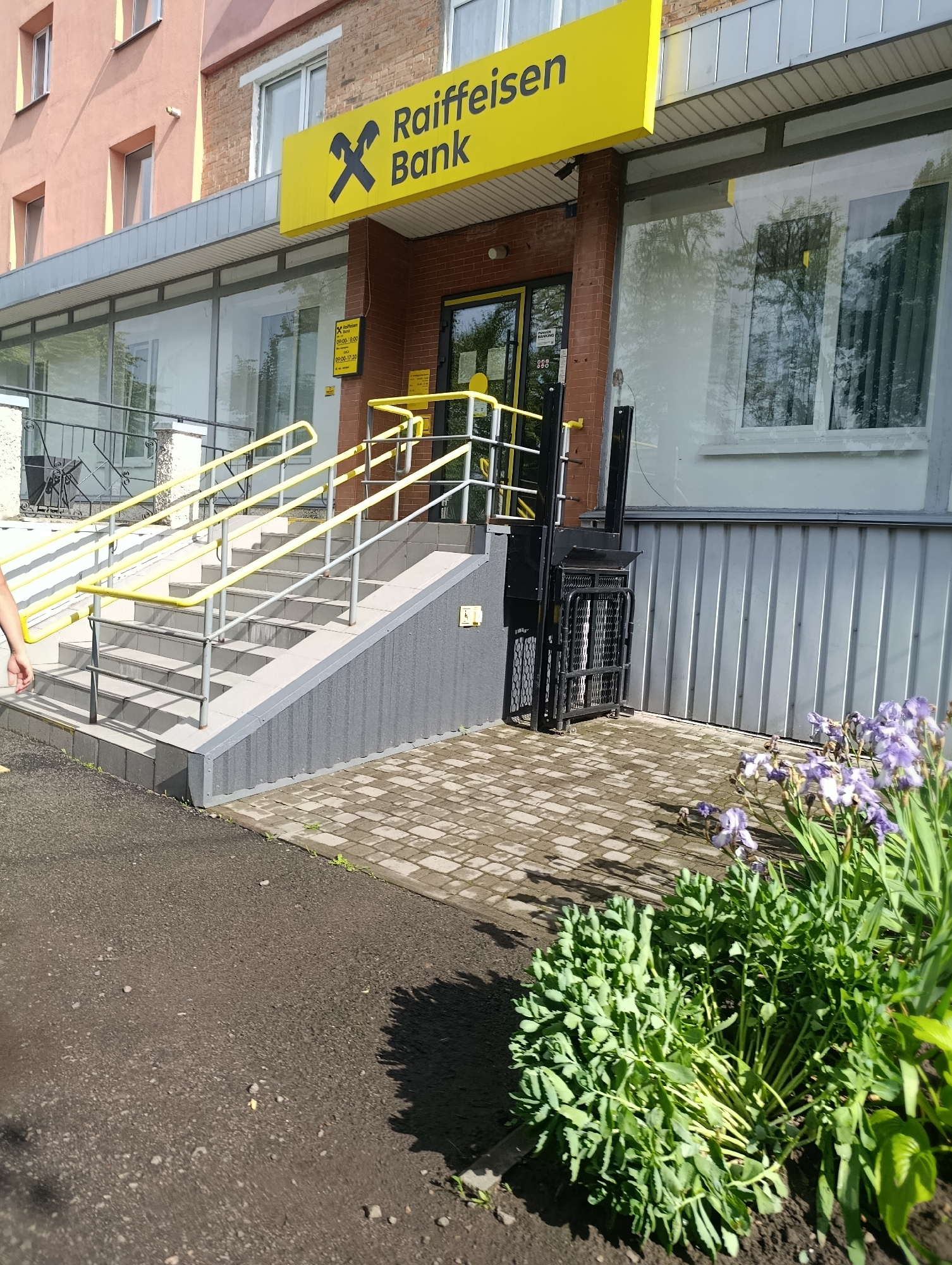 The image shows the exterior of a Raiffeisen Bank branch in Khorol, Ukraine. In the foreground, there is a paved area with some green foliage and purple irises. To the left, a dark asphalt surface is visible. A set of outdoor stairs with yellow railings leads up to the entrance of the bank. To the right of the stairs, there is a wheelchair lift. The bank itself has large glass windows and a yellow sign with the Raiffeisen Bank logo and text. The building is made of brick and has a light-colored facade. The overall scene depicts a typical urban bank entrance on a sunny day.