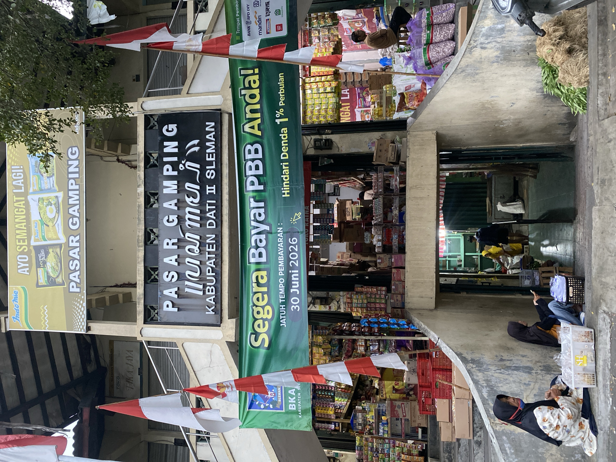The image depicts the entrance to Pasar Gamping, a market located in Ambarket বৈশিষ্ট্য, Kabupaten Sleman, Indonesia. The market entrance is framed by concrete structures and stairs. A large green banner with yellow text reads "Segera Bayar PBB Anda! JATUH TEMPO PEMBAYARAN: 30 Juni 2026" and "Hindari Denda 1% Perbulan," indicating a local tax payment deadline.
In the foreground, two women wearing headscarves are sitting on the concrete steps. The woman on the left, in a floral patterned dress, has a white basket containing plastic bags in front of her. The woman on the right, in a black and orange headscarf, is holding a mobile phone.
Above the entrance, a black sign with white text reads "PASAR GAMPING" followed by text in a script that appears to be Javanese, and then "KABUPATEN DATI II SLEMAN." Further up, a yellow banner features the "Indomie" logo and the text "AYO SEMANGAT LAGI!". Indonesian flags, red and white, are prominently displayed on poles.
The market stalls are visible behind the banner, filled with a variety of packaged goods, including food items and what appear to be cleaning supplies. A man is seated on a stool to the right, observing the scene. In the background, another person is walking through the market. To the far right, a motorcycle is parked near a pile of green vegetables and straw. The overall environment suggests a bustling local market scene with commercial activity and community interaction.