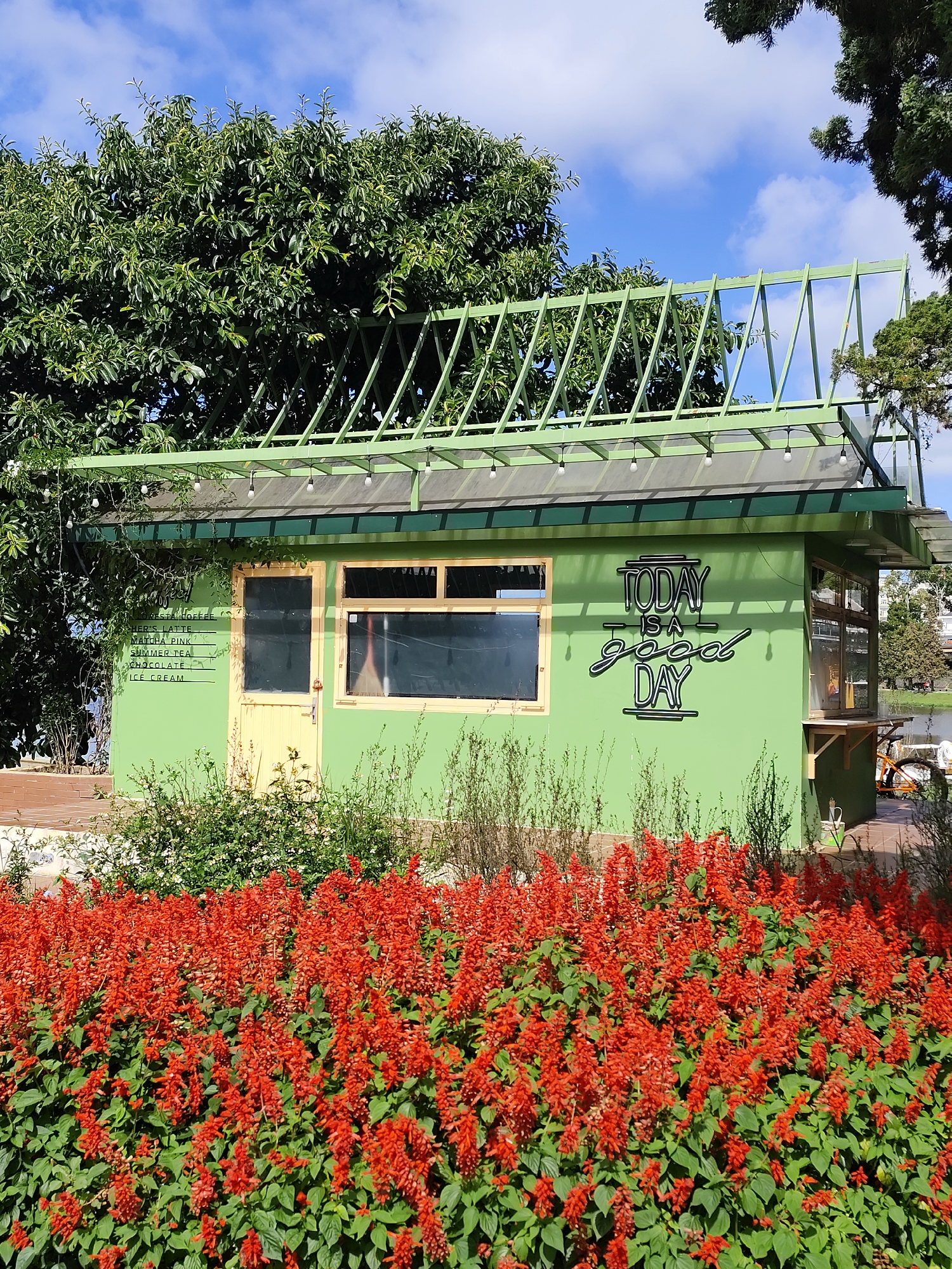 The image depicts a green, one-story building located in Da Lat, Vietnam. A sign reading "TODAY IS A good DAY" is attached to the side of the building. Another sign listing "RESTRO COFFEE, MATCHA PINK, SUMMER TEA, CHOCOLATE, ICE CREAM" is attached to the front of the building. The building is surrounded by bright red salvia flowers. Above the building is a lattice structure partially obscured by trees. The sky is visible in the background.
