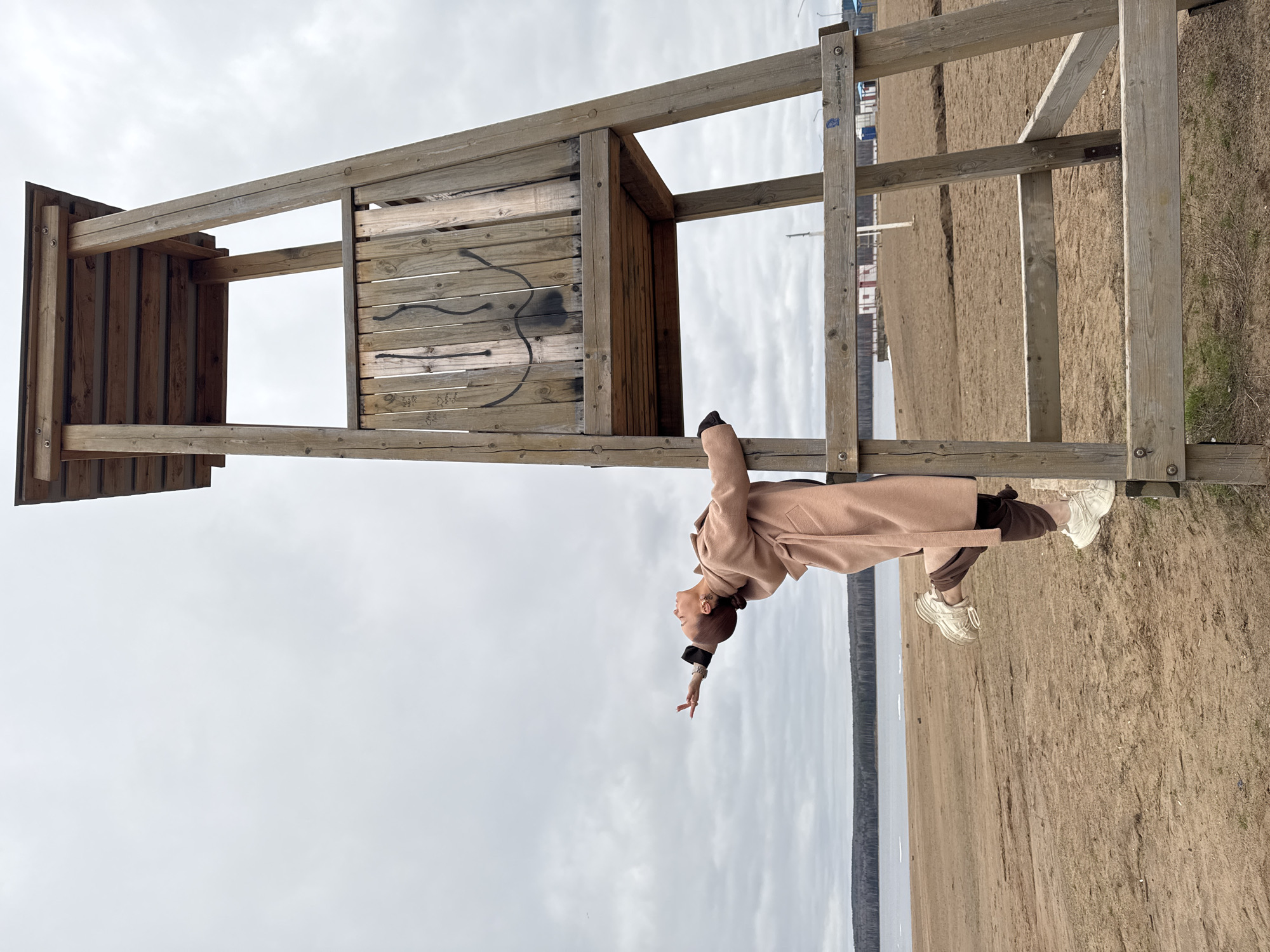 An inverted full-shot photograph captures a young woman performing an acrobatic pose on a weathered wooden structure set on a sandy shoreline under an overcast sky. The woman, positioned centrally within the frame, appears to be hanging upside down from a horizontal beam of the structure. She is dressed in a long, light brown coat, dark trousers, and light-colored chunky sneakers. Her head is tilted back, and her right arm is extended downwards with fingers splayed, creating a dynamic and stylized posture. Her hair is pulled back, revealing earrings and subtle details around her neck.
The large wooden structure, likely a lifeguard stand or observation platform, is constructed from sturdy, light brown planks and beams, showing signs of weathering and age. On the rear, enclosed wall of the structure, faint dark graffiti, including a prominent wavy line and some indistinct alphanumeric characters, is visible.
The scene unfolds on a wide, sandy beach or shore, which occupies the lower portion of the image and stretches into the midground, featuring sparse patches of dry grass. Beyond the sand, a calm body of water extends to a distant horizon, met by a dense, dark treeline. The sky is uniformly cloudy and light gray, suggesting an overcast daytime without direct sunlight. The overall atmosphere is quiet and somewhat muted, consistent with a cool or mild day. The location context is Chaikovsky, Russia, fitting for a large body of water such as the Kama River or Votkinsk Reservoir.