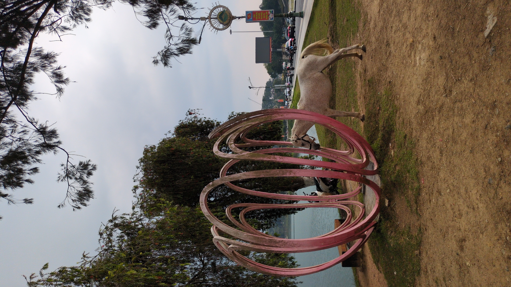 An outdoor scene features a white horse sculpture in front of a lake. The horse is positioned to the right of a large, pink, abstract spiral structure. In the background is a road with vehicular traffic and buildings. Trees are on the left side of the image. The location appears to be in Da Lat, Vietnam.