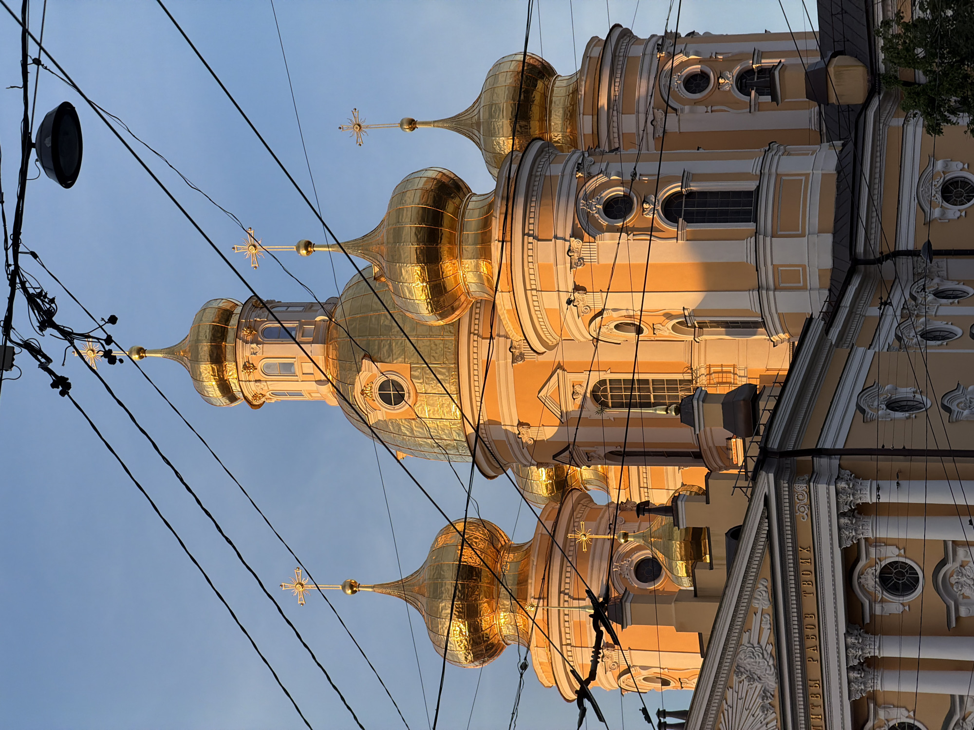 The image displays a majestic Orthodox church with gleaming golden domes set against a clear blue sky, partially obstructed by numerous overhead electrical wires. The building, likely located in Saint Petersburg, Russia, features multiple onion-shaped domes, each topped with an ornate golden Orthodox cross that includes sunburst-like rays.
The church's facade is primarily a light, creamy yellow or peach color, adorned with white architectural details such as pilasters, cornices, and decorative moldings, suggesting a Baroque or Neoclassical style. Various windows are visible, including arched and circular designs, some with intricate grilles. The warm, golden light illuminating parts of the structure indicates either late afternoon or early evening, creating a striking contrast with the shaded areas.
A dense network of black electrical wires and cables crisscrosses the foreground, partially obscuring the view of the church and the sky. A dark street light or speaker is also visible on the left, connected to these wires. A hint of green foliage from a tree can be seen on the far right.
On a decorative frieze of the church, visible in one of the provided crops, text in Old Church Slavonic reads: "МОЛИТВЫ РАБОВ ТВОИХЪ". This translates to "Prayers of Your servants." The sky is clear, suggesting fair weather.