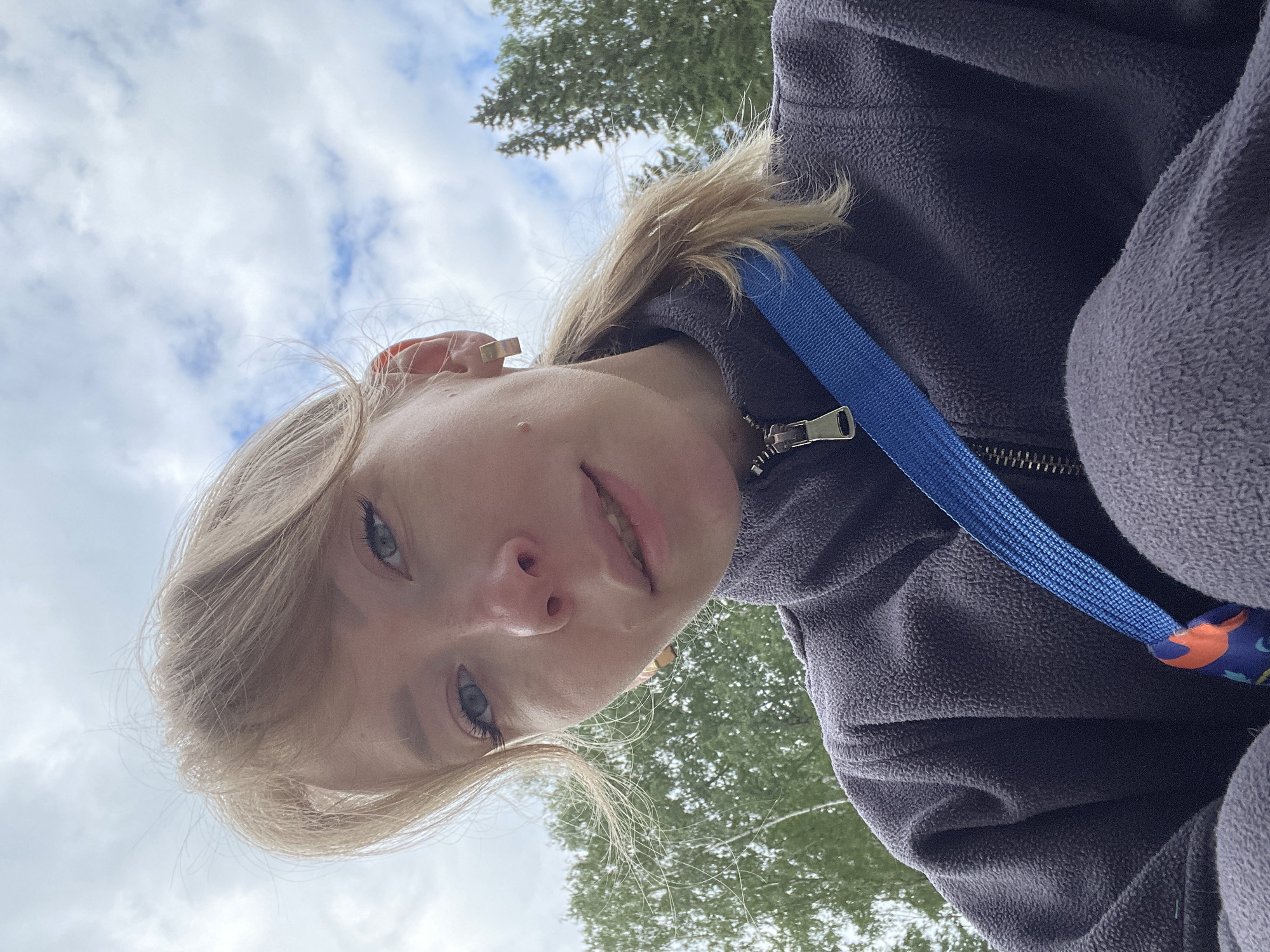 The image is a low-angle selfie of a young person, likely female, looking directly into the camera from an upward perspective. The subject has fair skin, light-colored blonde or light brown hair, and striking blue eyes with visible mascara or eyeliner. A small mole is noticeable on her left cheek (viewer's right). She wears a dark grey or purple fleece jacket with a visible zipper and a bright blue lanyard or strap draped across her chest. A small, light-colored earring is visible in her right ear. Her expression is calm and relaxed, with a faint smile.
The setting appears to be outdoors during the daytime, under a bright, cloudy sky with patches of blue. In the background, green trees, possibly evergreens, suggest a natural environment such as a park or forest, consistent with the location context of Perm, Russia. The unique angle of the shot implies the photo was taken while the subject was looking up, possibly lying down. There is no visible text in the image.