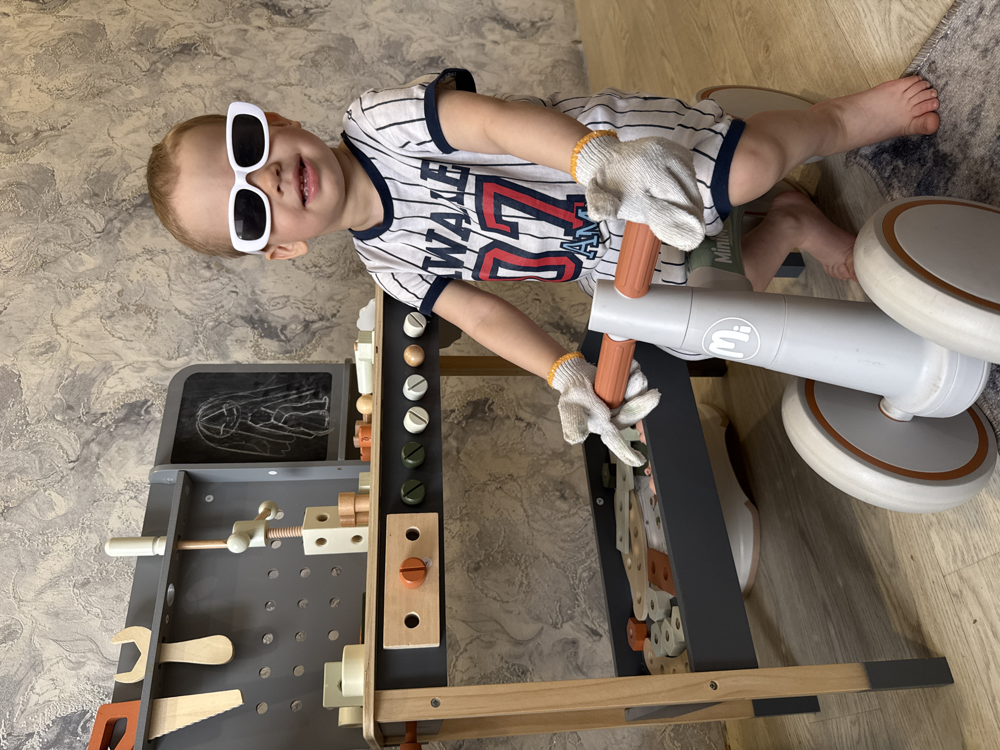 A young child is depicted in an indoor setting, smiling brightly while wearing white-framed sunglasses and white work gloves with orange stitching. They are dressed in a white and black striped t-shirt featuring the text "WAKE 27 AWAY" in red and black.
The child is positioned near a gray and wooden toy workbench, which is equipped with various wooden tools and parts, including a vise, screws, nuts, bolts, a wrench, and a saw. A small chalkboard on the workbench displays a simple chalk drawing. Adjacent to the child and the workbench is a gray and white ride-on toy or scooter, distinguished by orange accents on its handles and wheel rims. The ride-on toy features a circular "Mii" logo and the word "Mini" printed on its side. The child is holding onto the handles of this toy, suggesting active play or using it as part of their engagement with the workbench.
The scene is well-lit, indicating it is daytime. The background features a marbled gray and brown textured wallpaper and a light wood laminate floor. A small patterned rug is visible beneath the ride-on toy. The child's happy expression and playful attire suggest a moment of joyful and imaginative play.