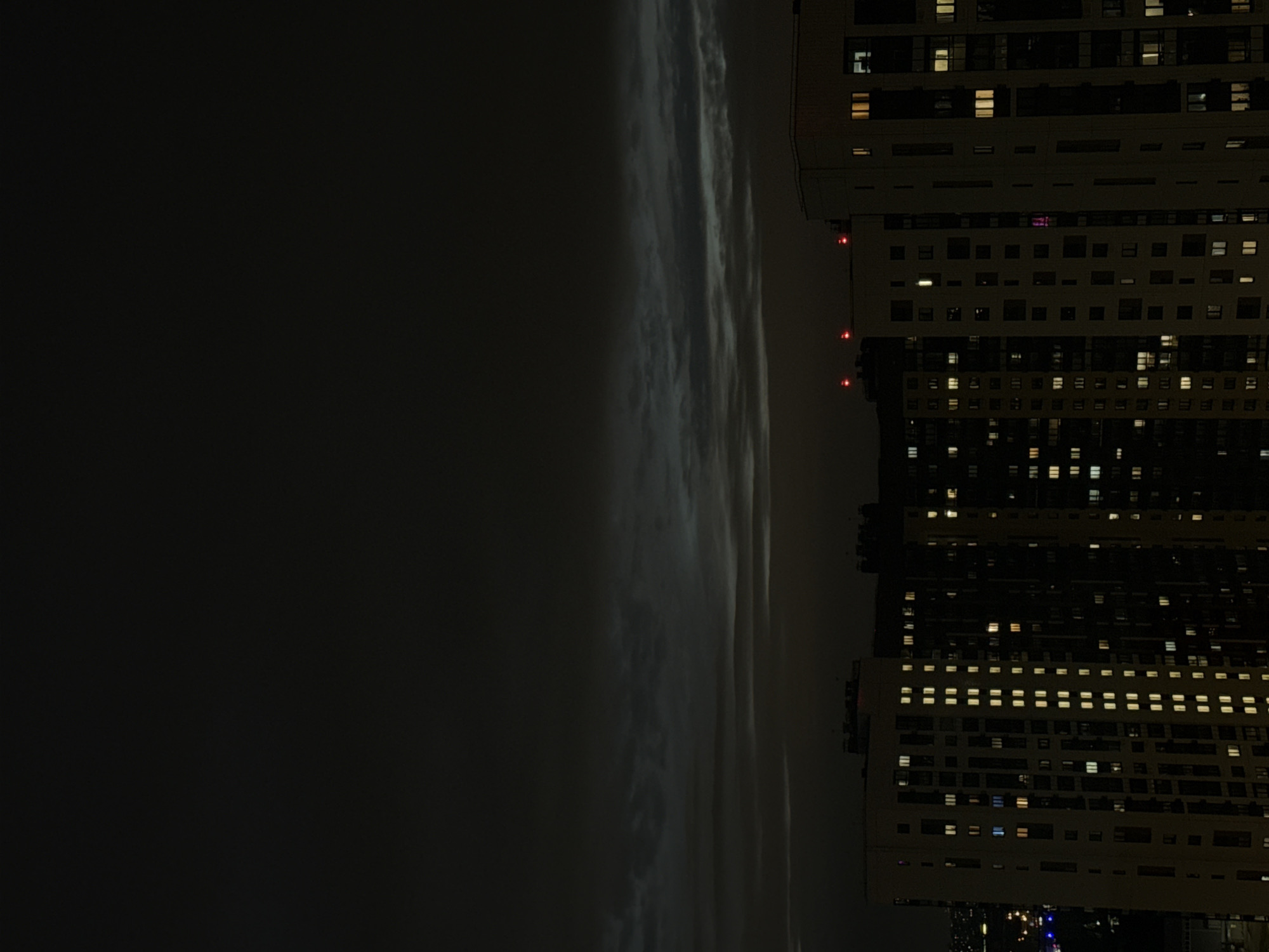 The image shows a nighttime view in the city of Lyubertsy, Russia. The right side of the frame features a tall apartment building, illuminated with numerous lights in its windows. Three red lights are visible on the upper portion of the building. To the left of the building, a dark sky is visible, partially obscured by what appears to be fog or low-hanging clouds. The image is oriented with the sky and building appearing sideways.