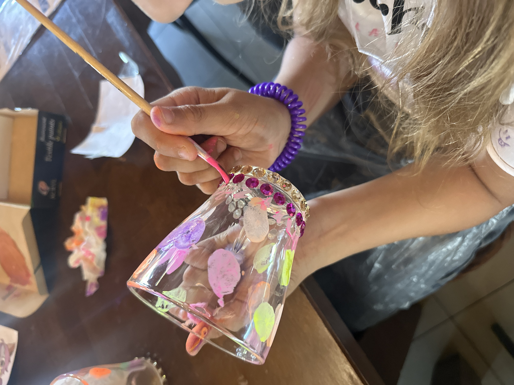 A fair-skinned individual is painting a clear glass cup adorned with gemstones around the rim. The cup is decorated with various colorful paint dabs in shades of pink, purple, orange, and green. The individual wears a purple coil bracelet on their left wrist and holds the cup with their left hand, while the right hand holds a paintbrush with pink paint on the bristles. The scene takes place on a brown wooden table with various items nearby, including a box and pieces of tissue. The location is Unknown, Unknown.