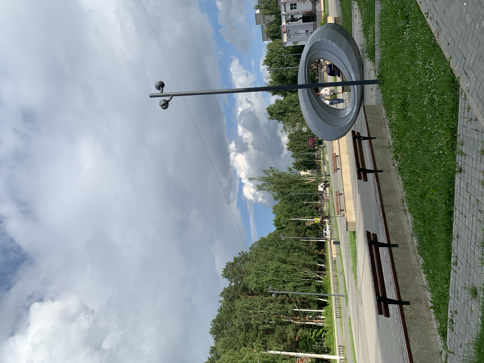 The image captures an outdoor scene in the city of Chaikovsky, Russia, specifically in a park-like area with a street and buildings in the background. In the foreground, there are several wooden benches with concrete supports, set against a backdrop of lush green grass and a paved walkway. To the right, a large, abstract metallic sculpture with an oval opening is prominently displayed. Two individuals are partially visible within the sculpture's opening, and another person is seated on a bench behind it. A tall lamppost with two fixtures stands next to the sculpture. In the midground, a road is visible with a few cars and pedestrians. The background is dominated by a dense line of tall green trees, likely a forest or park. The sky is partly cloudy, with large white cumulus clouds against a blue sky. The overall impression is of a public recreational space on a bright, overcast day.