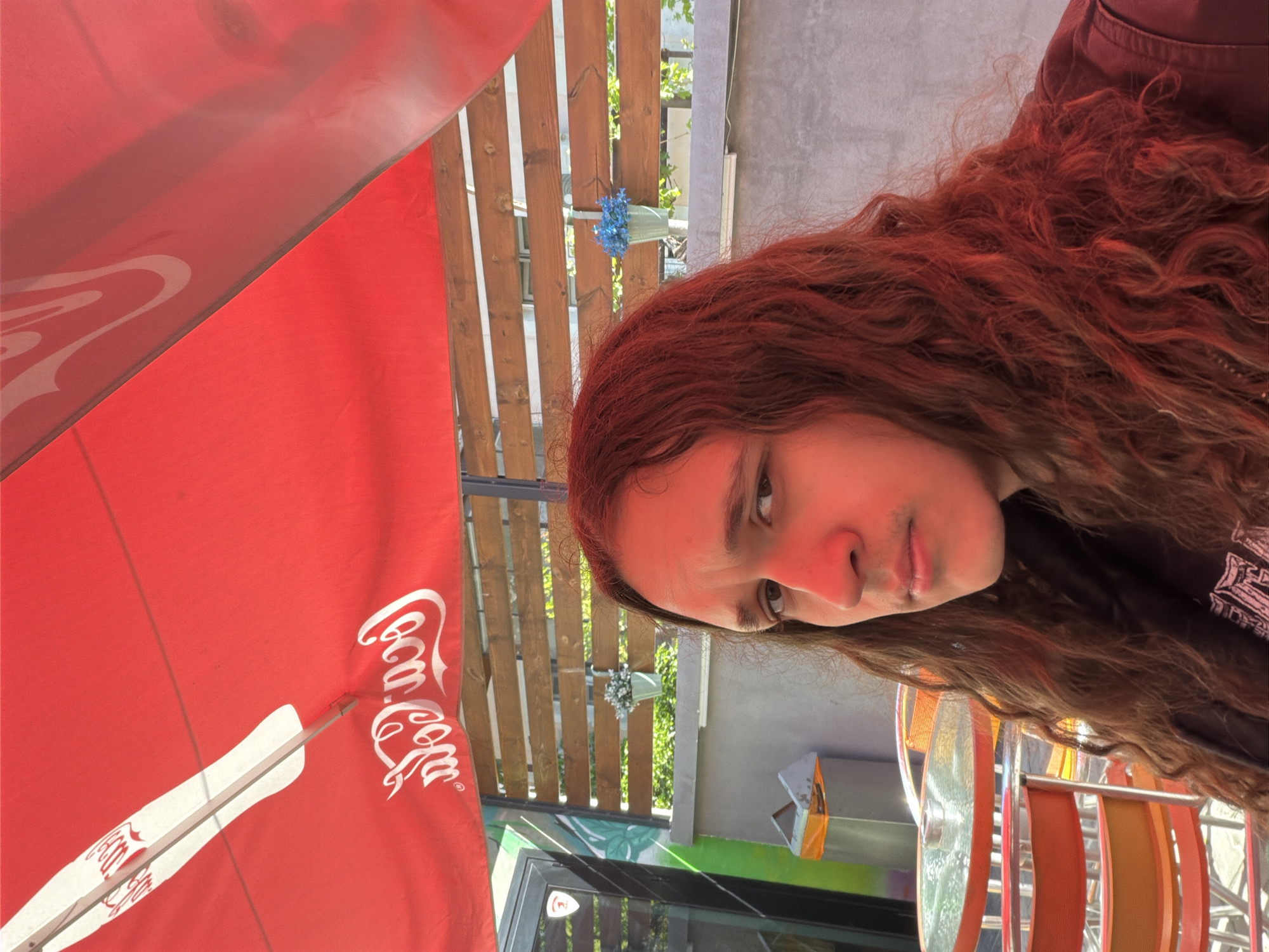 A close-up, slightly tilted, medium-shot captures a fair-skinned individual with long, wavy, auburn hair. The person is gazing towards the left, with a slight frown on their face. Part of their body is visible, clad in a dark-colored garment.
In the backdrop, a red Coca-Cola-branded umbrella is partially visible, casting some shade. The presence of a light-colored wooden fence and a small blue flower arrangement attached to it suggests an outdoor setting, potentially a cafe or restaurant terrace. Additionally, there is an outdoor seating area with colored chairs.
The location is likely in Plovdiv, Bulgaria, based on the style of the architecture and the type of umbrella used.