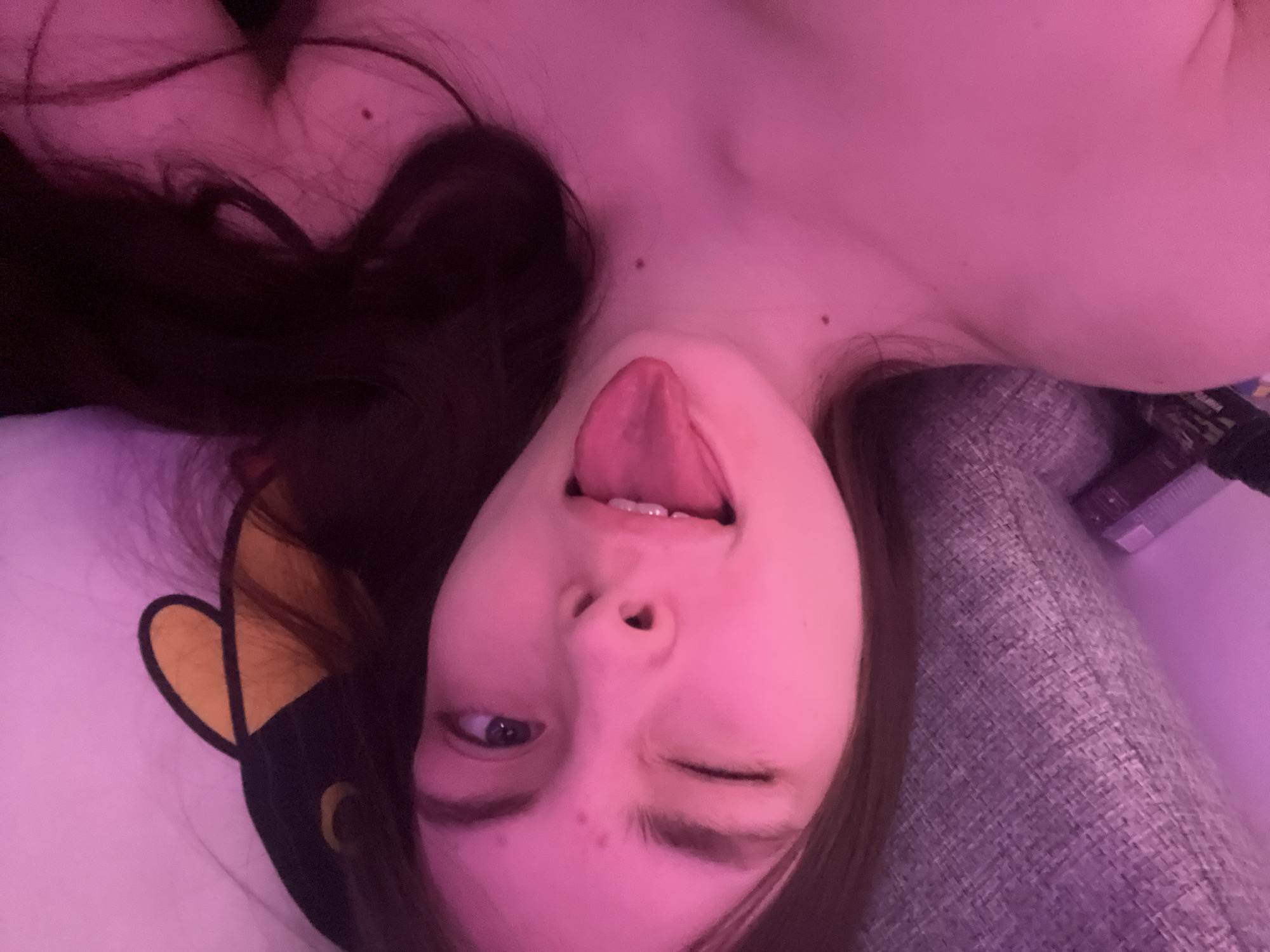 The image is a close-up selfie of a young woman with long, dark hair. She is lying down, possibly on a bed or couch, with her head tilted to the side. She is sticking out her tongue and winking. The lighting is pink-toned. A sleep mask is visible to the left, positioned above her head. The background is blurry and indistinct, and there is no indication of being located in the city of Saint Petersburg, Russia.