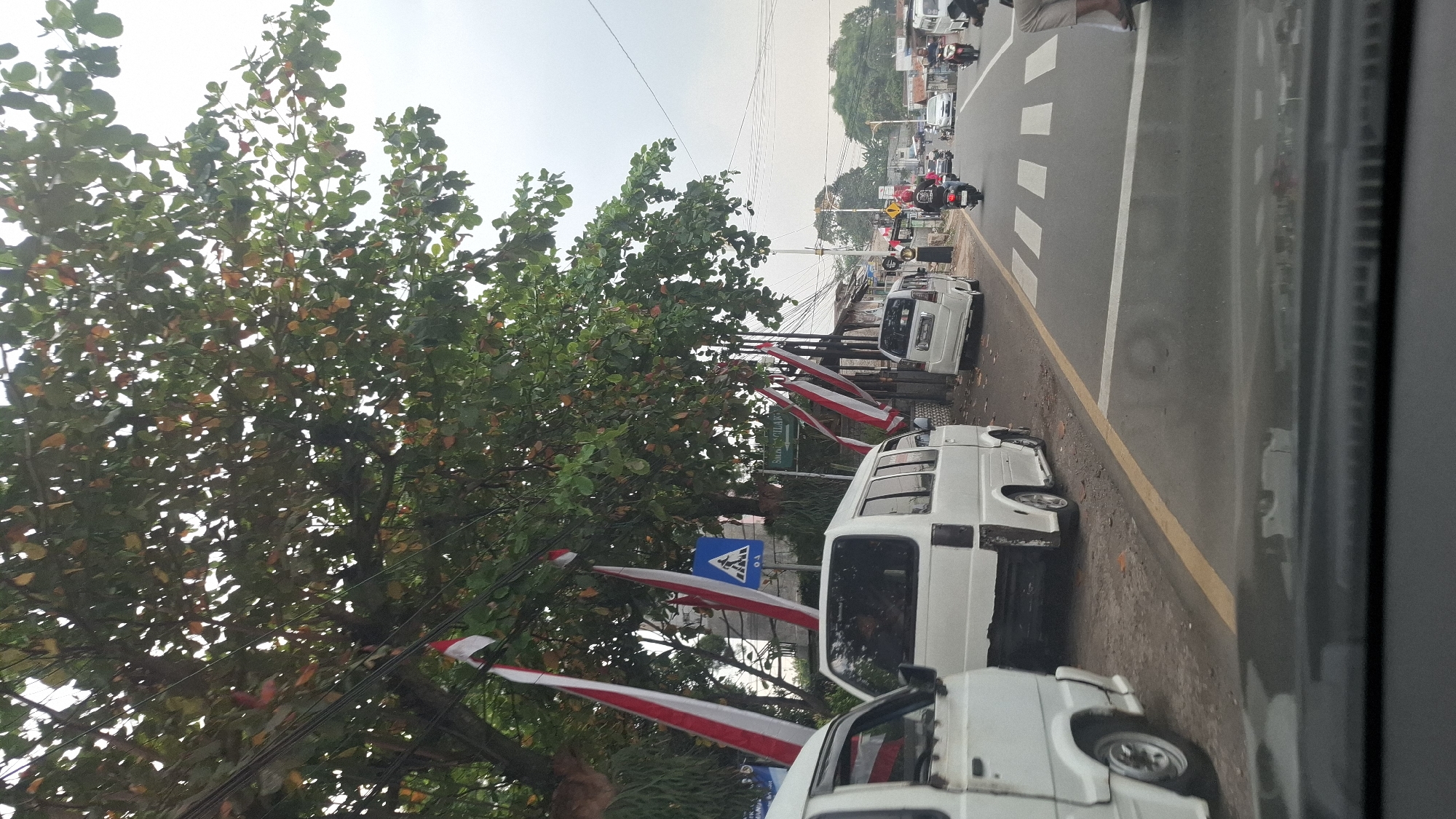 The image depicts a street scene in Cilaku, Indonesia, under an overcast sky. The perspective is from inside a vehicle, looking forward. The foreground is dominated by the dashboard and windshield of the vehicle.
In the midground, a two-lane road stretches into the distance. Several white minivans are parked or driving along the side of the road. One minivan is positioned on the left, partially visible. Another is parked on the left shoulder, with its rear facing the camera. Further down the road, another white minivan is in motion. A motorcycle with a rider is also visible on the right lane, heading away from the camera.
Flanking the road on the left are trees with green and reddish-brown leaves, and several Indonesian flags are hung vertically from wires and poles, their red and white stripes visible. A blue pedestrian crossing sign with a white symbol is mounted on a pole amongst the trees. A green directional sign with white lettering reads "SUMUR CICILAK."
The background shows buildings with awnings and various structures lining the road, suggesting a commercial or residential area. The overall impression is a typical street view in an Indonesian town.