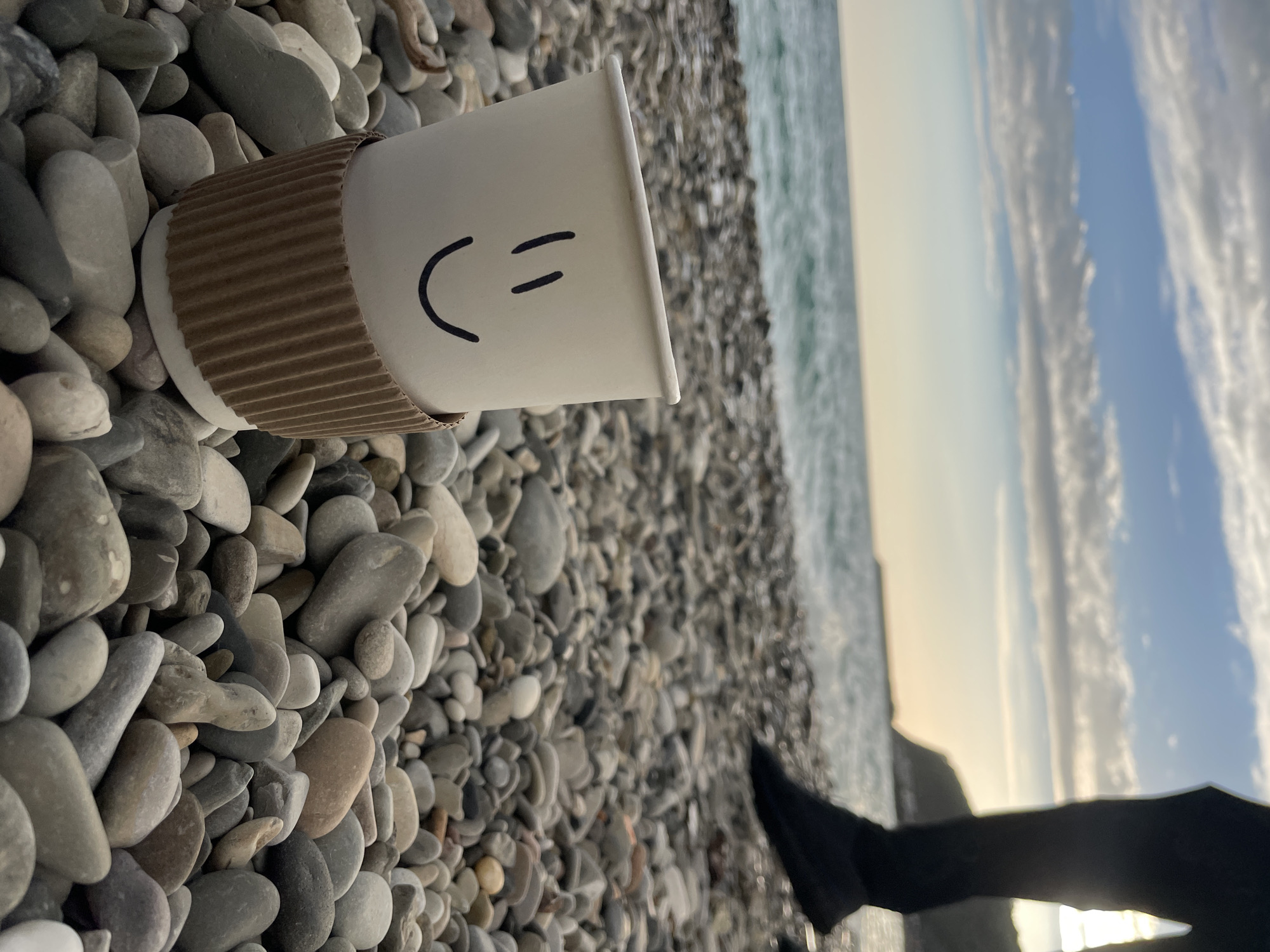 A white disposable coffee cup with a cardboard sleeve and a smiley face drawn on it sits on a beach of pebbles. The sea is in the background, and the sky is partly cloudy. A dark object, possibly a person's leg in black pants, is visible in the lower right corner. The location appears to be a beach in Olginka, Russia, based on the provided details.