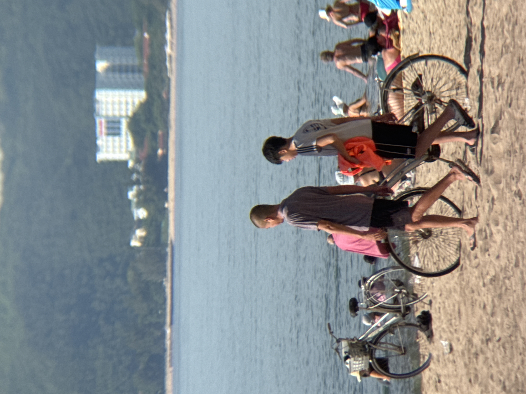 Two young men walk on a sandy beach in Nha Trang, Vietnam, with bicycles. One man, wearing a grey t-shirt and black shorts, carries a bright orange life vest. The other, in a grey t-shirt and dark shorts, walks beside him. Behind them, the calm blue ocean stretches to the horizon, where a hazy shoreline is visible with a large, light-colored building complex nestled among trees. Several other beachgoers are scattered on the sand and in the water. The lighting suggests a sunny day.