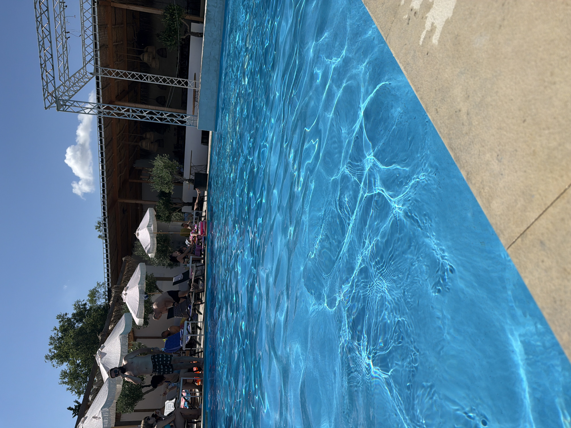 The image depicts a lively outdoor scene by a swimming pool, likely in Burgas, Bulgaria, given the context. A group of people are relaxing under white umbrellas and on lounge chairs, some are in swimwear. One man with a beard, wearing patterned swim trunks, stands near a lounge chair, holding a cigarette. Another man in swim trunks is bent over a lounge chair. The pool itself is a vibrant blue, with sunlight creating shimmering patterns on its surface. In the background, a covered structure with thatched roofing and hanging lamps suggests a restaurant or bar area. A metal scaffolding structure is visible to the right, indicating a possible entertainment or event setup. The environment is bright and sunny, with a clear blue sky and some clouds. The presence of olive trees adds a touch of Mediterranean ambiance. The overall atmosphere appears relaxed and recreational.