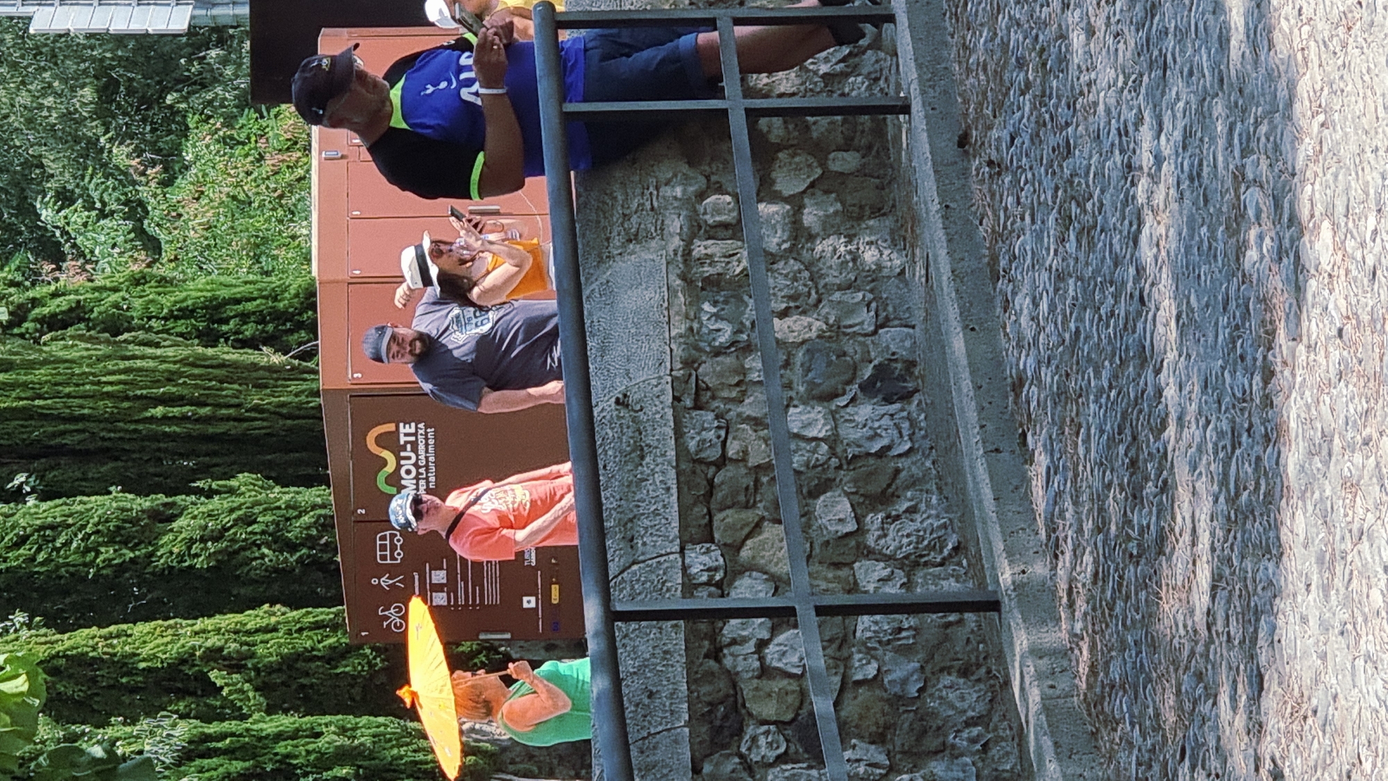 In the city of Besalú, Spain, a group of people stand along a stone wall. On the left, a woman in a green top holds an orange umbrella, shielding herself from the sun. Next to her, a man in an orange t-shirt and a baseball cap looks to his left. To his right, a man in a dark t-shirt and baseball cap is looking down at his phone. Beside him, a woman wearing a white hat and an orange top is also looking at her phone. On the far right, a man in a blue t-shirt and a baseball cap is looking down at his phone. Behind the group, a brown structure with signage is visible, featuring icons of a bicycle, walking figures, and a bus. The background is filled with tall green trees. The foreground shows a cobblestone path.