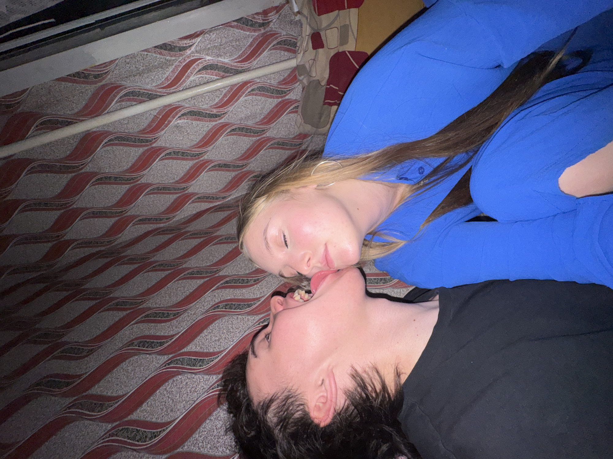 The image shows two young people close together against a backdrop of patterned curtains. The person on the left, with dark, tousled hair, is wearing a black t-shirt and has their mouth open with their tongue extended towards the other person. The person on the right, with blonde hair and closed eyes, is wearing a blue collared shirt. They are positioned in a way that suggests an intimate interaction. The environment appears to be indoors, with a curtain featuring a wavy red and gray pattern. No specific details of the city of Perm, Russia are visible in the image.
