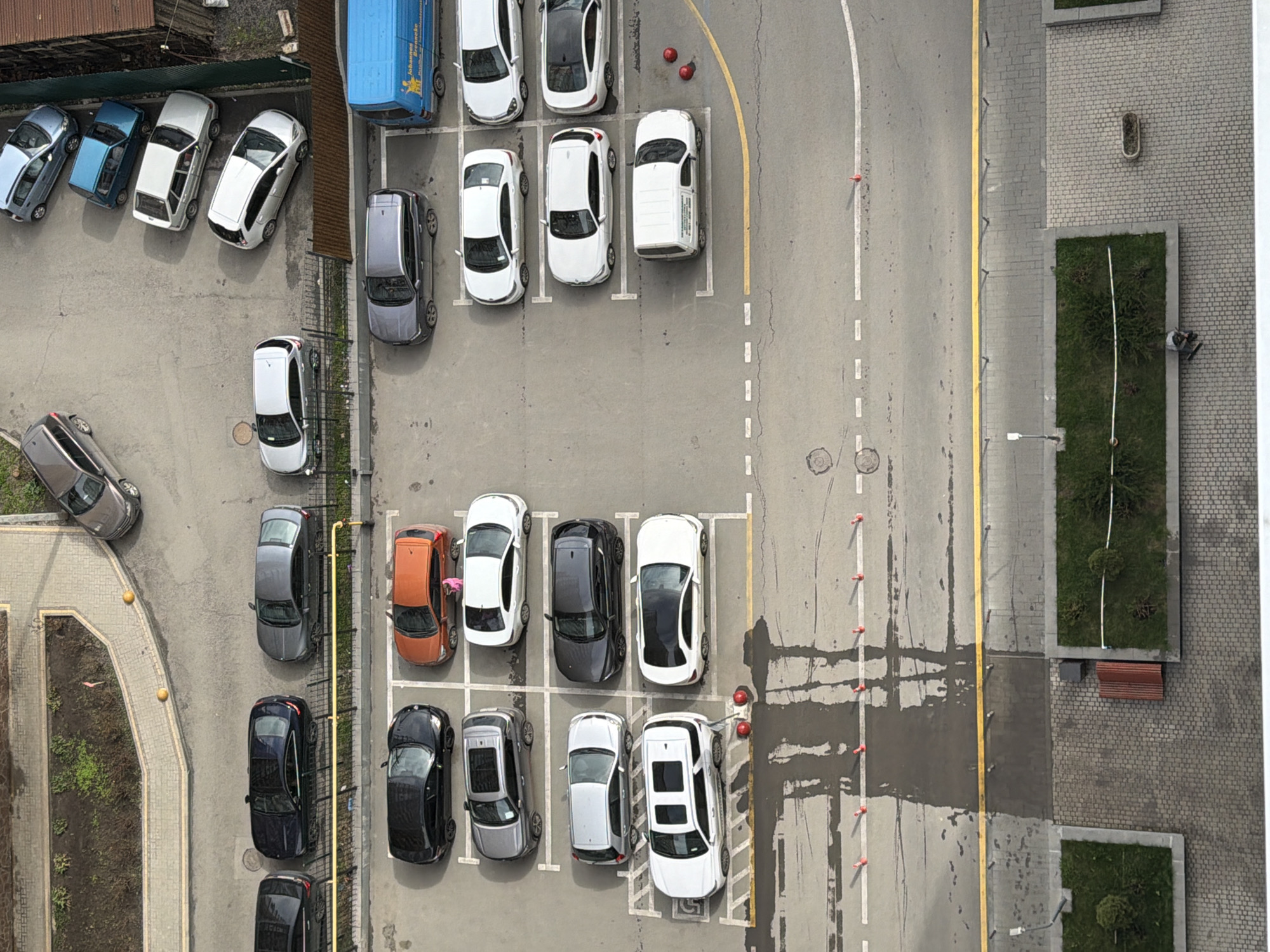 The image is an aerial view of a parking lot in what is presumed to be the city of Azov, Russia. The parking lot is mostly filled with cars of varying colors and sizes. There are parking spaces marked with white lines, and a yellow line designates a lane. The parking area appears to be adjacent to a building with green spaces including trees and a small park area. There are no people clearly visible in the image. There is no visible interaction happening in the scene.