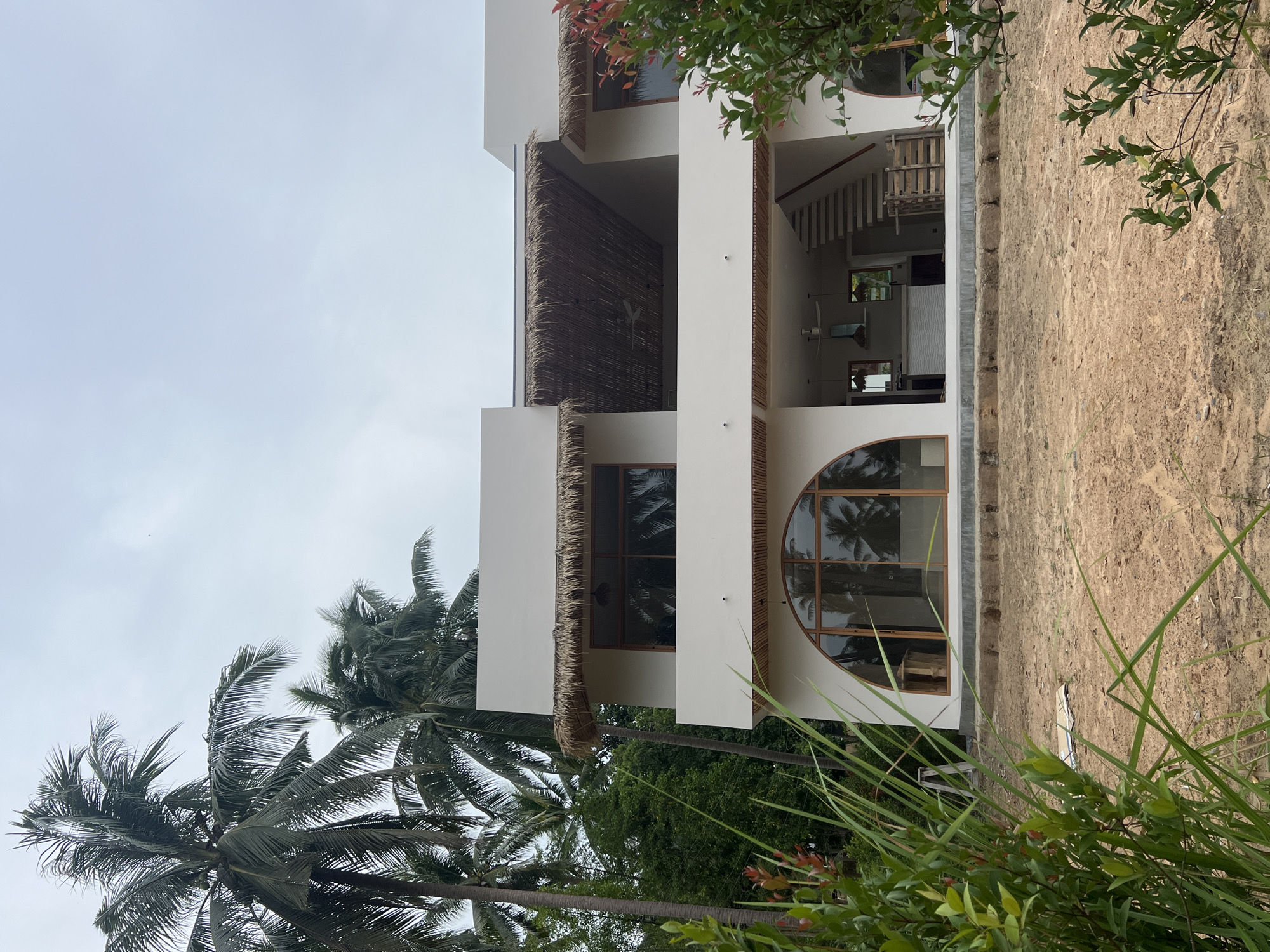 The image shows a two-story building under construction, situated amidst palm trees on a plot of sandy land. The building has a modern design with white walls and large windows, some of which are arched. The roof has a thatched overhang. Inside, visible through the lower level windows, are remnants of construction materials, including what appear to be kitchen cabinets and a staircase. The foreground is dominated by lush green vegetation and tall grasses. The sky is overcast. The location is identified as the Ko Pha-ngan District in Thailand.