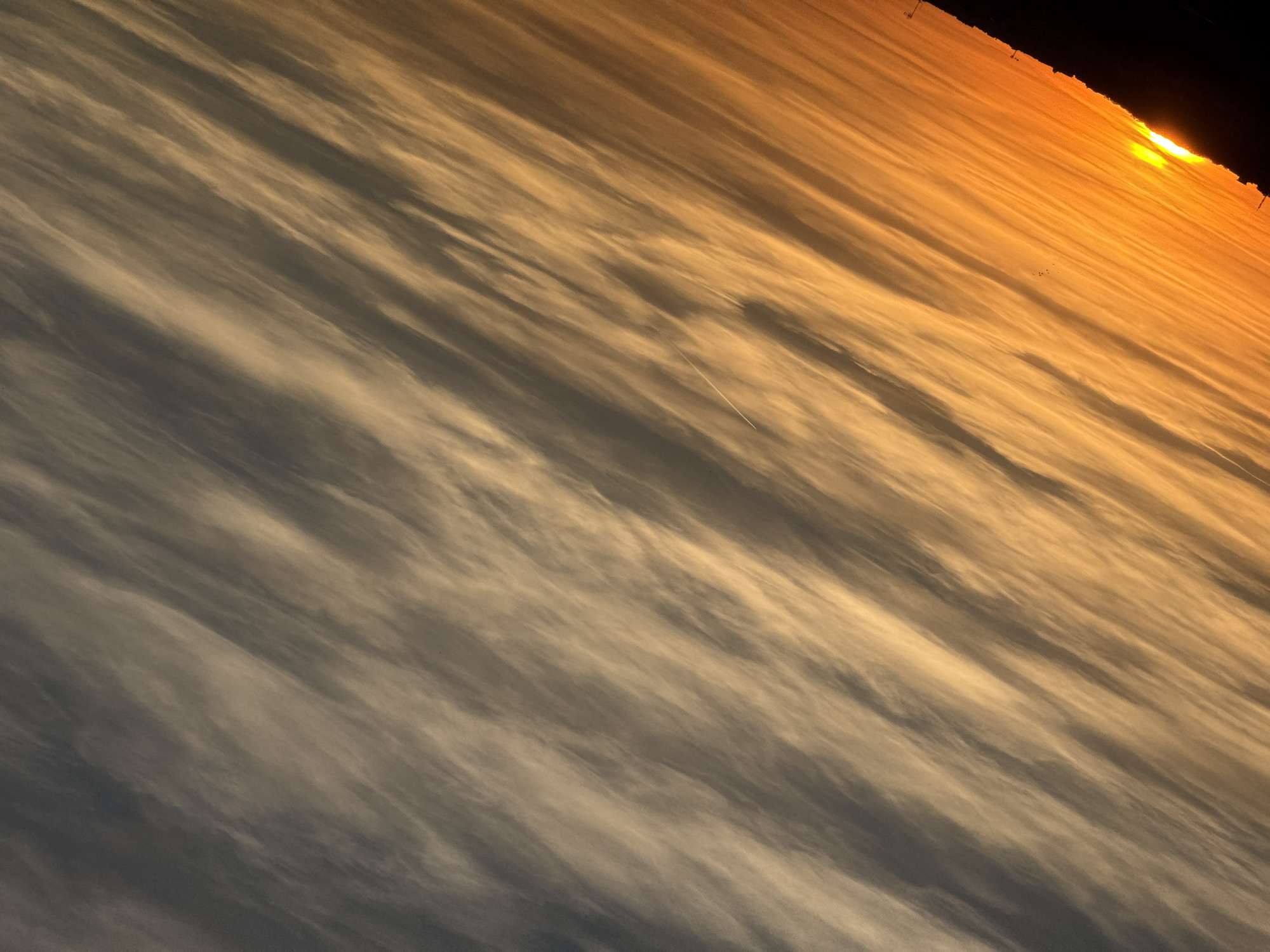 This is a photograph of the sky taken at an upward angle, possibly from an airplane or a high vantage point. The image is dominated by clouds, which are streaked and wispy, suggesting they are moving rapidly. The lighting indicates that it is either sunrise or sunset, with the sun visible as a bright, glowing orb on the right side of the frame, casting a warm, orange glow across the clouds. The sun appears to be low on the horizon, and its light is diffused by the atmospheric conditions.
The clouds themselves are a mix of dark gray and lighter, almost white, tones, creating a dramatic contrast. The streaks in the clouds are oriented diagonally across the frame, adding a sense of motion and depth. In the upper right portion of the image, a dark silhouette of what appears to be structures or buildings can be seen against the bright sky. There are also a few tiny dark specks that could be birds or other small aircraft, and a thin white contrail left by an airplane is visible in the middle of the frame. The overall impression is one of dynamic atmospheric conditions and the beauty of a sky at dawn or dusk. The image evokes a sense of awe and wonder at the natural world. No text is visible in the image.