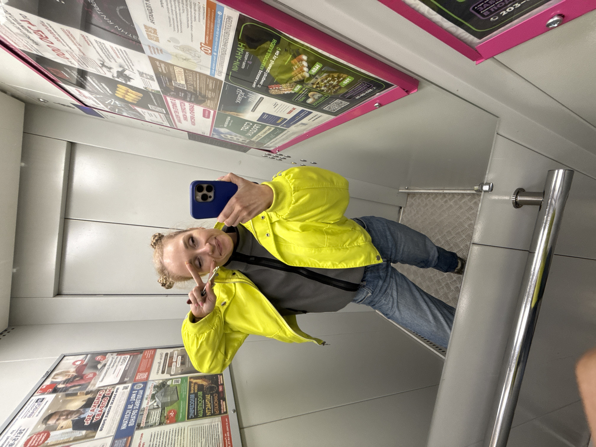 The image captures a young woman taking a mirror selfie in what appears to be an elevator or a narrow, enclosed public space in Perm, Russia. The image itself is rotated, making the subject and environment appear tilted.
The central subject is a light-skinned woman with blonde hair styled in two small buns on top of her head. She is smiling broadly and playfully making a peace sign with her right hand, while holding a blue smartphone with a triple-lens camera setup in her left hand, presumably to take the picture. She is wearing a vibrant, neon yellow jacket over a grey long-sleeved top and blue jeans.
The setting consists of light grey walls. To the left and behind her, the walls are covered with various advertisements and posters, many framed in a distinctive magenta/pink color. These posters feature Cyrillic text, images of food (like sushi/rolls and pizza), people, and various promotional offers. Some visible text includes "НОВЫЙ ДОСТУП" (New Access), "АКЦИЯ" (Sale/Promotion), "БЕЗЛИМИТ" (Unlimited), "УДОБНО" (Convenient), and "ВЫГОДНО" (Profitable), along with phone numbers and brand names like "NPN-AHAH." A metal handrail is visible on the right side of the frame, and a section of textured metal flooring is seen below her legs. The overall impression is of a public transport interior or an elevator cabin. The time of day is not discernible as the space is artificially lit.