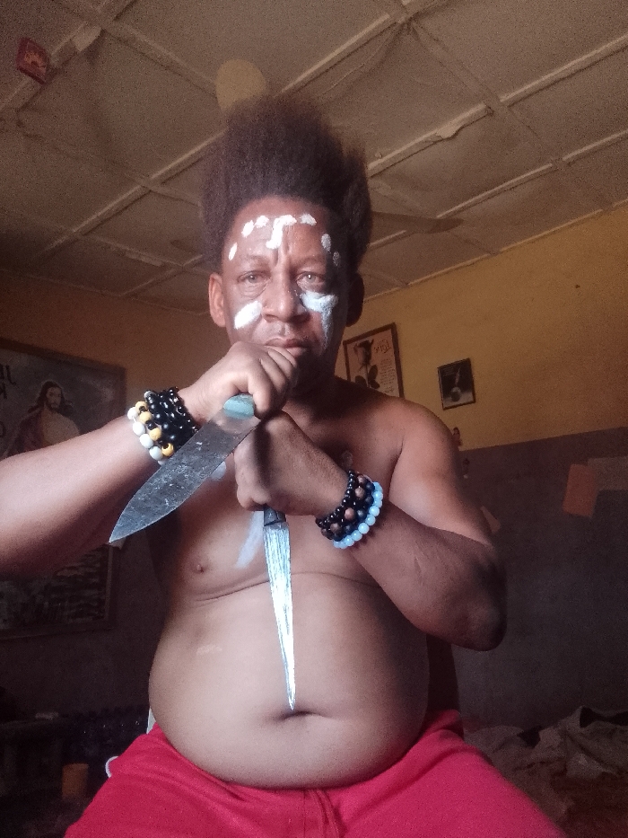An adult male is depicted in a medium shot indoors, identified as being in Narayi, Nigeria. The subject, bare-chested with a visible abdomen, has dark skin and dark, voluminous hair styled upwards. His face is marked with white pigment on the forehead, forming a central "T" shape with scattered dots, and on the cheeks, along with a light blue streak below his right eye. He wears multiple beaded bracelets on both wrists: the left wrist has black, white, and yellow beads, while the right wrist displays dark brown and light blue beads. The man holds two large, sharp knives with both hands crossed over his midsection. The knife in his left hand, with a light-colored handle, points diagonally upwards. The knife in his right hand, with a dark handle, points downwards. Both knife blades appear metallic and show signs of use. The background features light brown walls and a white paneled ceiling, where a ceiling fan is partially visible. A framed picture of Jesus Christ is visible on the left wall, and two other framed images are indistinctly present on the right wall.