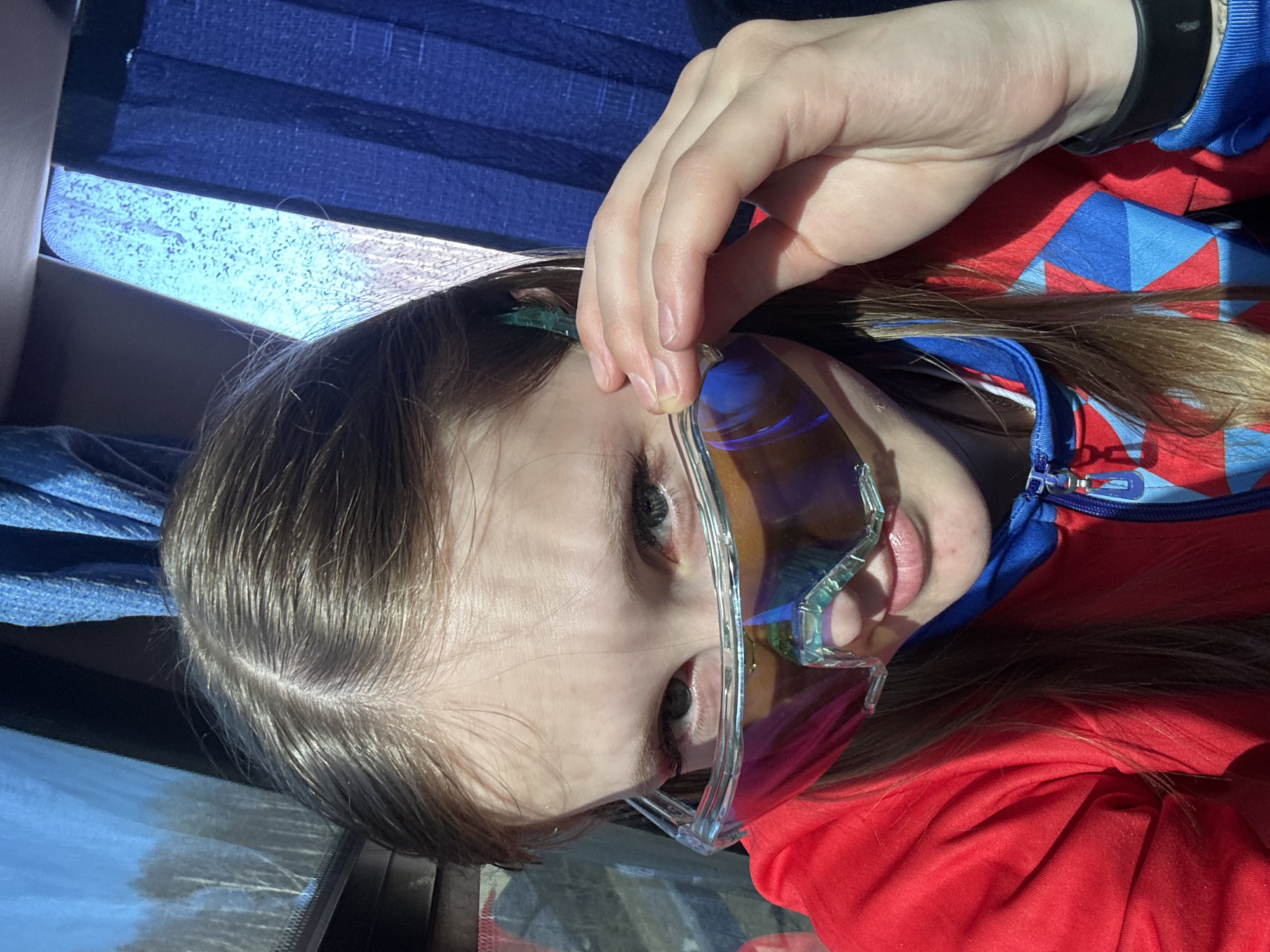 A person is holding a pair of iridescent sunglasses in front of their face. The individual has light skin, brown hair, and is wearing a red and blue jacket. The background includes a blue curtain and a window reflecting an outdoor scene. Although the scene appears to be taken in Berezniki, Russia, it is impossible to definitively confirm location based on the available image data.