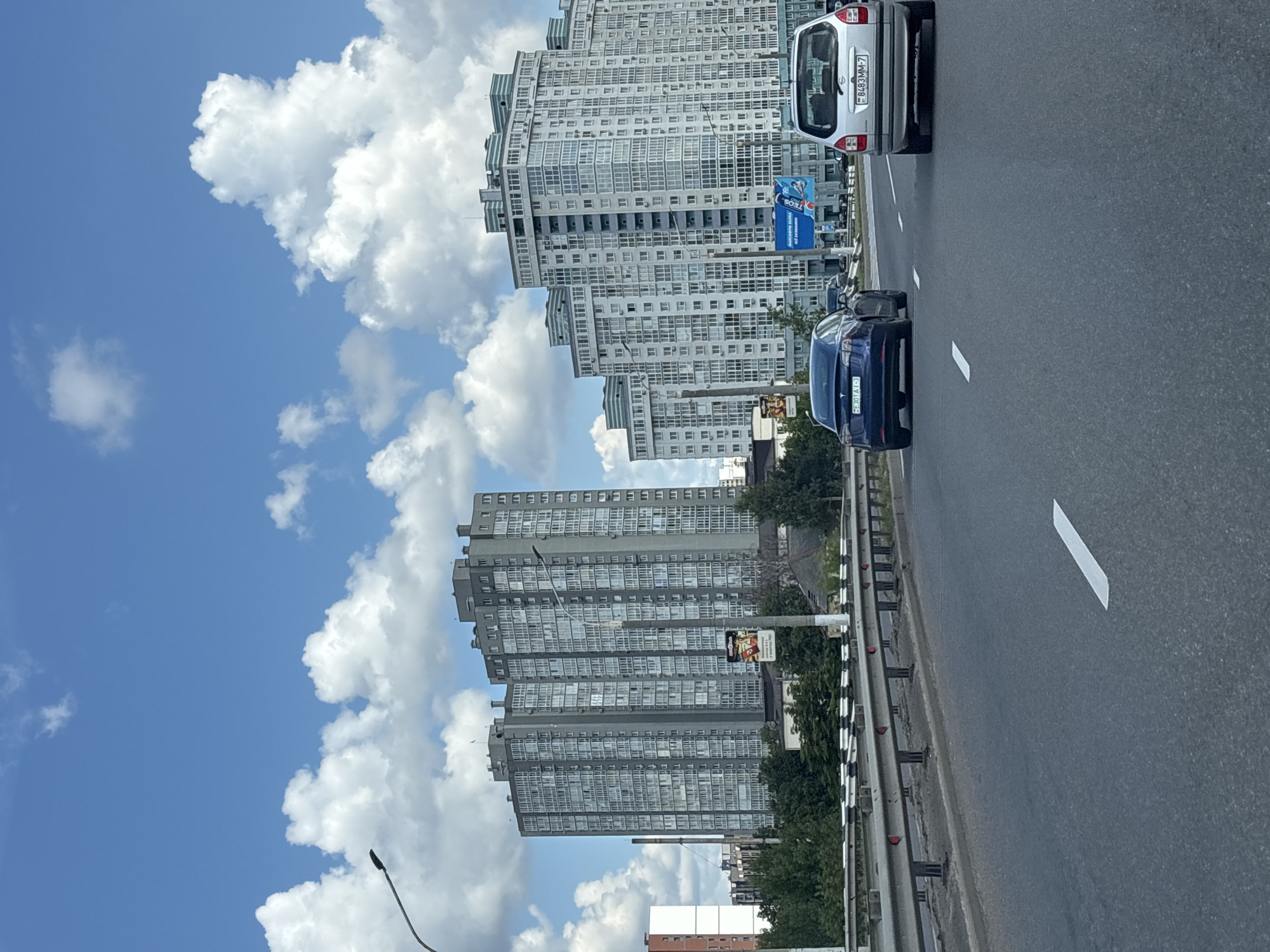 The image captures a view from a highway in Minsk, Belarus, featuring modern apartment buildings against a backdrop of a partly cloudy blue sky. Two multi-story residential buildings dominate the background, one a darker gray and the other a lighter color with distinctive turquoise accents on its upper floors. A highway with visible lane markings stretches into the foreground, with several cars driving on it. A blue Tesla is in the left lane, and a silver minivan is in the right lane. A metal guardrail separates the highway from an area with trees and smaller structures. Streetlights are visible along the highway. The overall scene suggests a daytime urban environment.